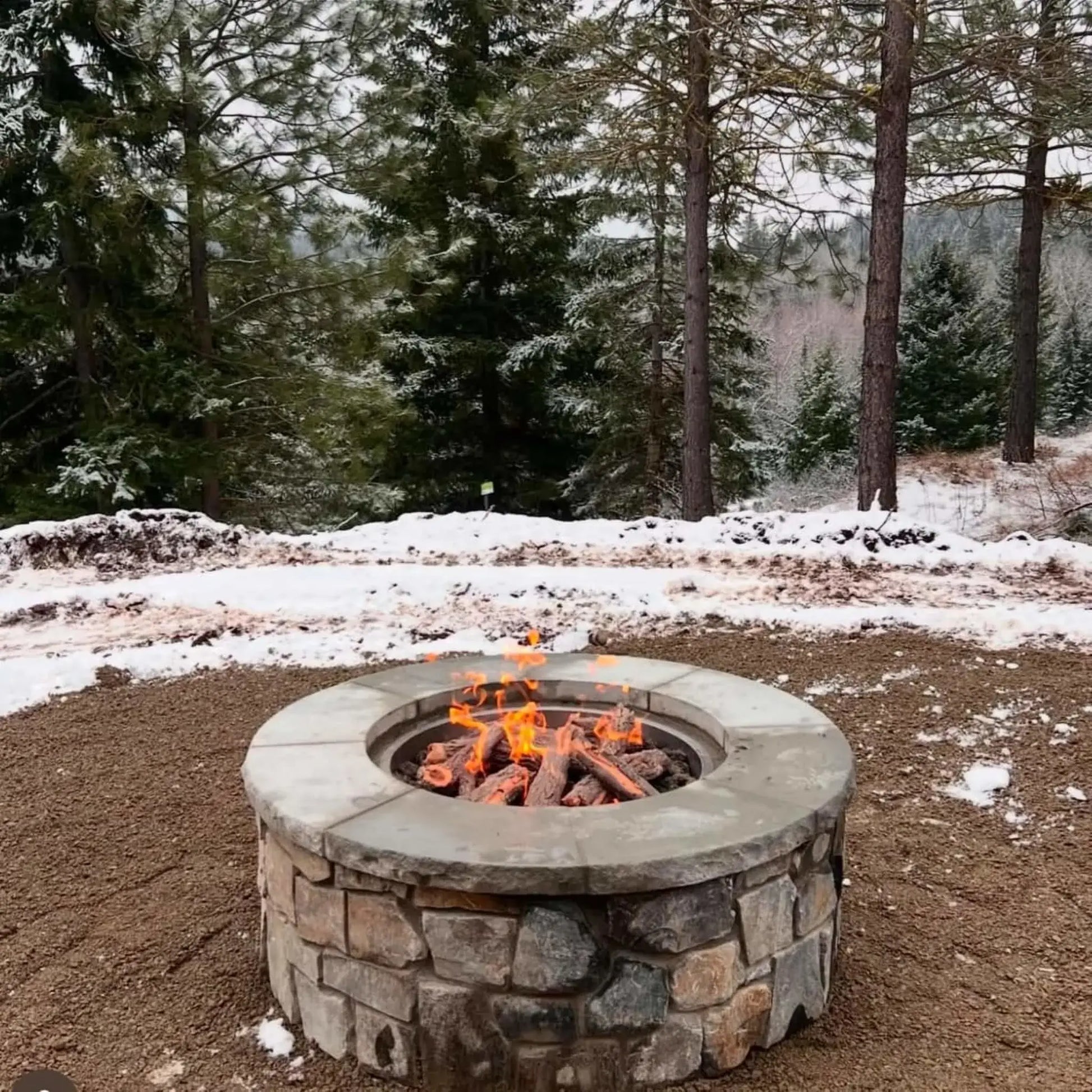 Round stone gas fire pit with weathered oak logs burning in a snowy forest setting. Surrounded by evergreen trees and light snow coverage, the glowing flames contrast against the cold environment, creating a warm and inviting winter outdoor fire feature.