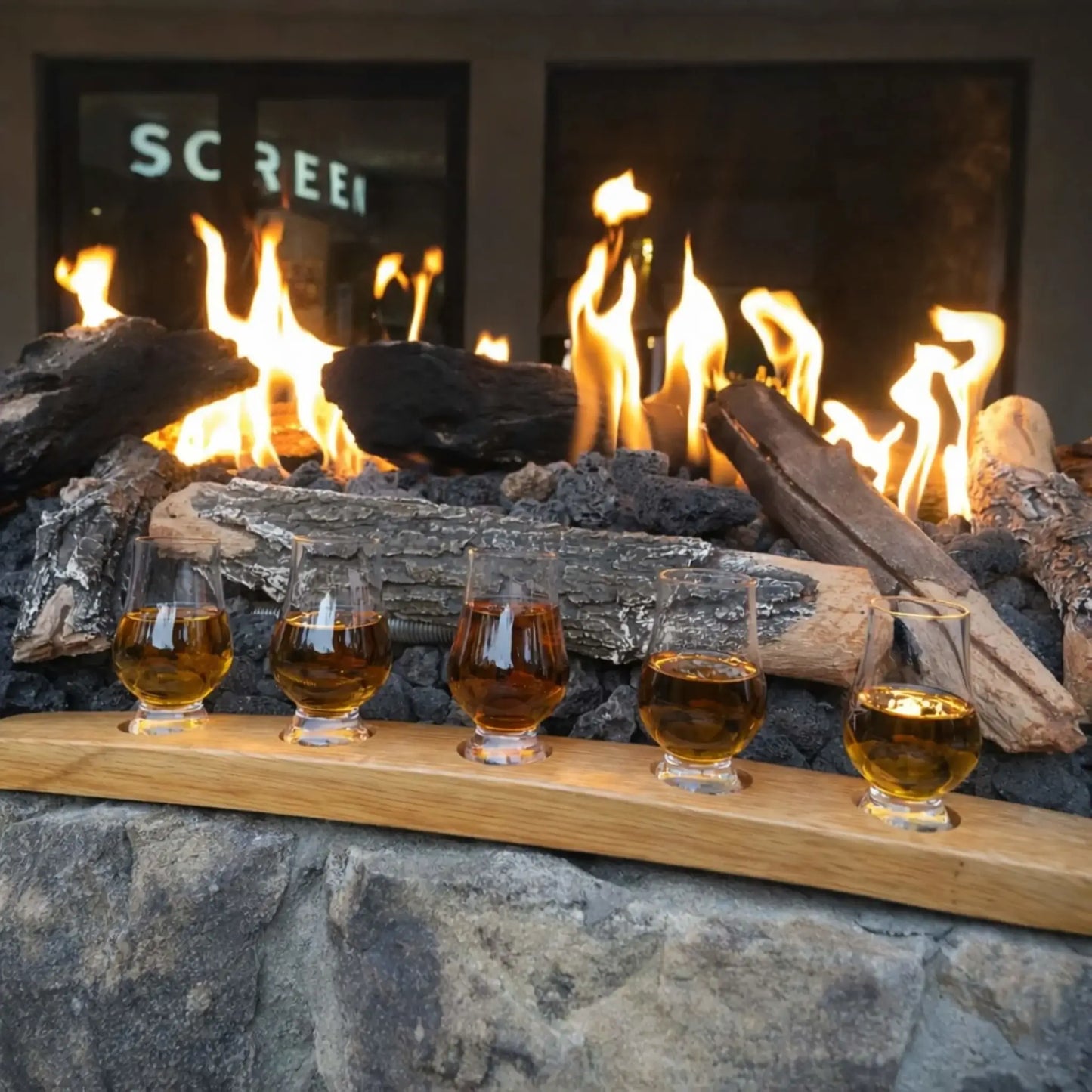 Close-up of a fire pit with weathered oak gas logs burning over dark lava rock, paired with a tasting flight of amber-colored drinks on a wooden tray in the foreground. The scene blends fire ambiance with a relaxed social setting, ideal for outdoor entertaining.