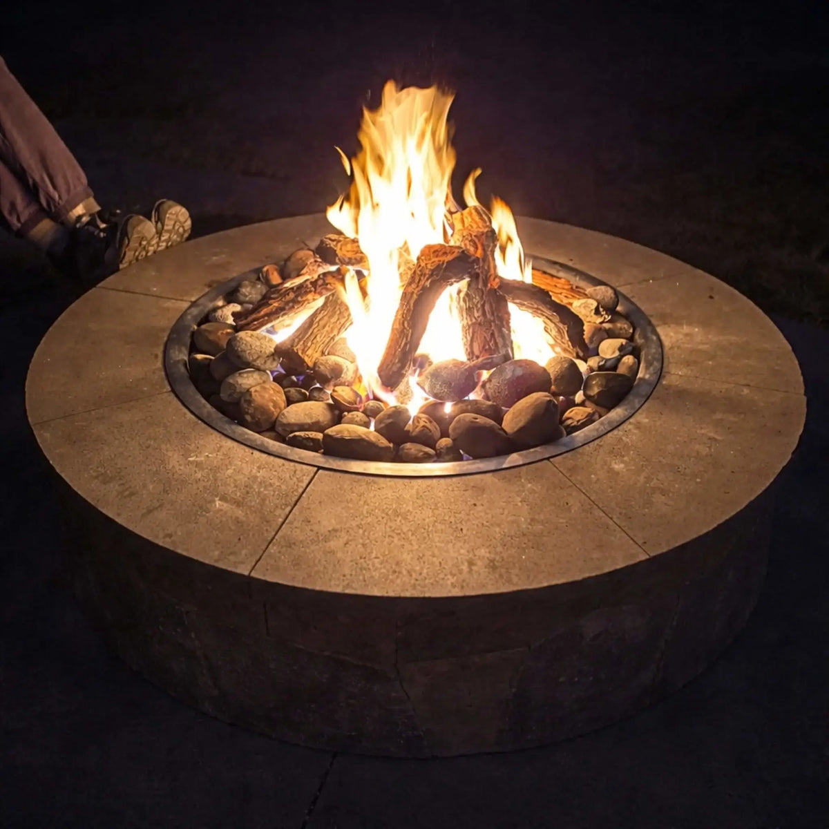 Evening view of a round stone fire pit with weathered oak gas logs arranged over river rock, producing bright flames against a dark background. A person’s legs rest nearby, emphasizing a relaxed, intimate atmosphere perfect for nighttime outdoor enjoyment.
