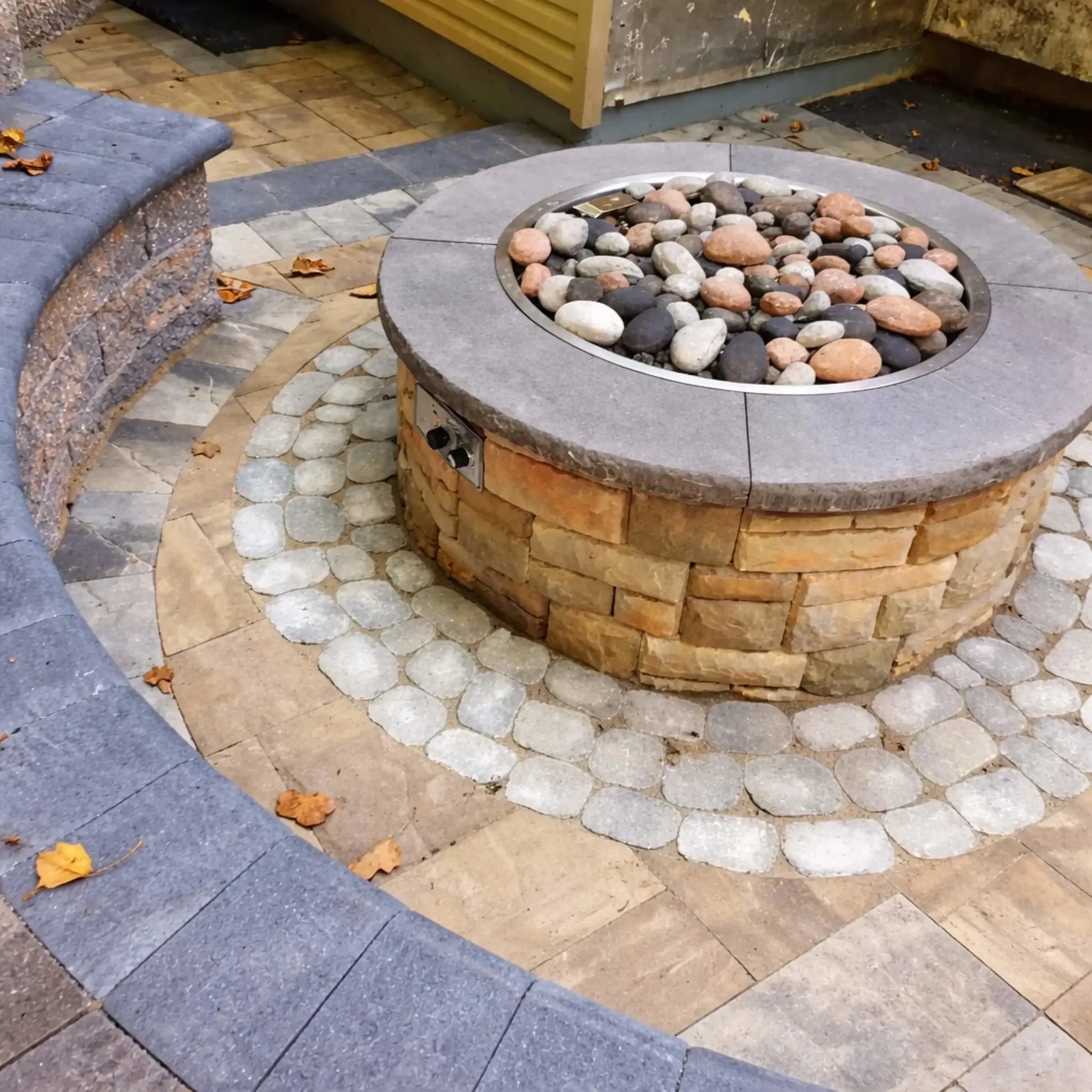 Circular gas fire pit built with stacked stone and a smooth concrete cap, filled with multicolor river rocks in gray, tan, and black tones. The fire pit sits within a patterned paver patio with a curved seating wall, highlighting a clean and inviting outdoor living space design.