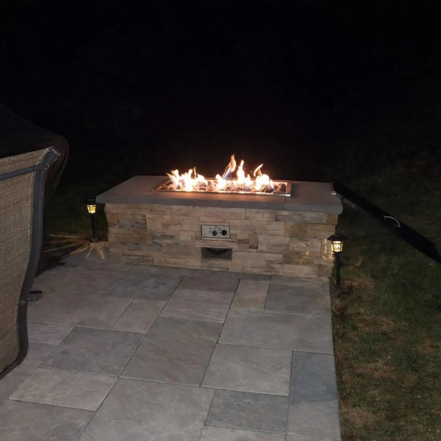 Rectangular outdoor gas fire table with a stacked stone base and stainless steel burner, filled with ceramic river rocks and active flames at night. The patio setting includes subtle landscape lighting and clean stone pavers, creating a modern and cozy backyard ambiance.