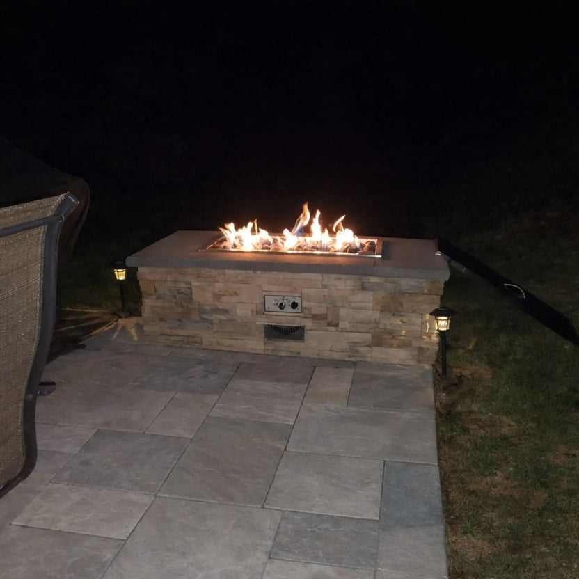 Rectangular outdoor gas fire table with a stacked stone base and stainless steel burner, filled with ceramic river rocks and active flames at night. The patio setting includes subtle landscape lighting and clean stone pavers, creating a modern and cozy backyard ambiance.
