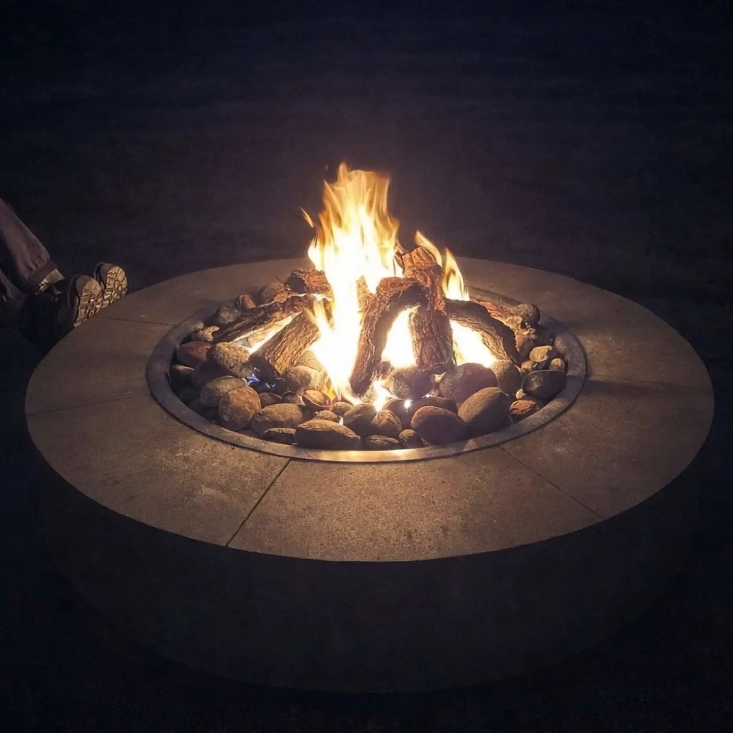 Round gas fire pit table at night with bright flames rising above a bed of smooth ceramic river rocks. The stones encircle realistic log pieces, while a person sits nearby, emphasizing warmth and comfort in a relaxed outdoor patio setting.