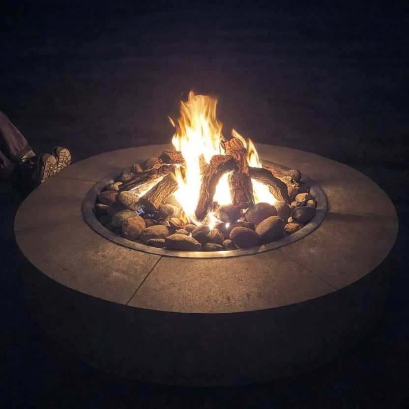 Round gas fire pit table at night with bright flames rising above a bed of smooth ceramic river rocks. The stones encircle realistic log pieces, while a person sits nearby, emphasizing warmth and comfort in a relaxed outdoor patio setting.