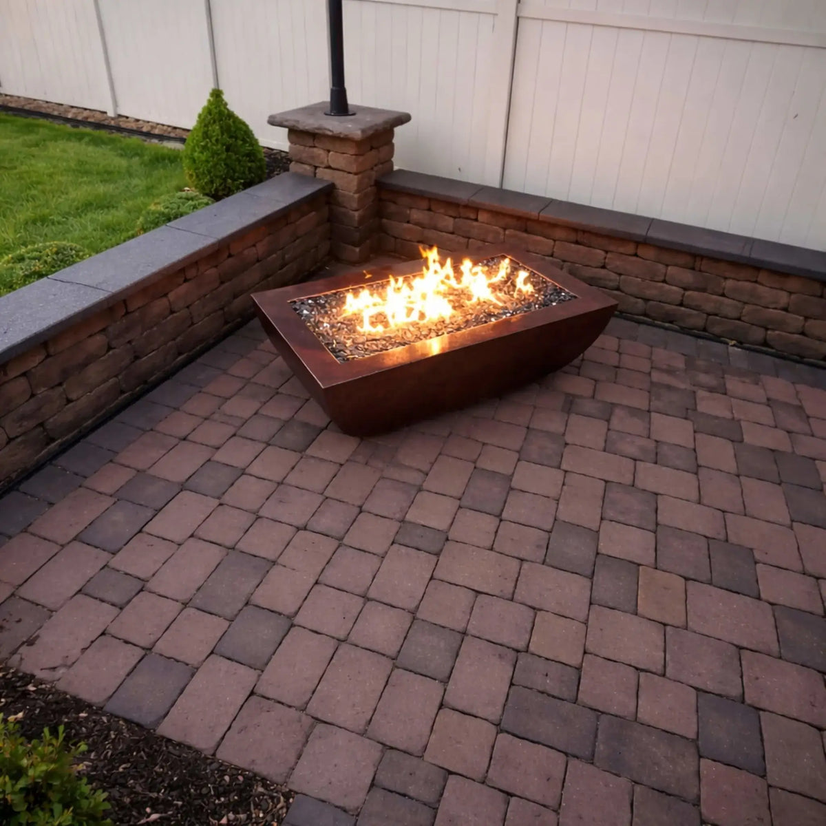 Backyard patio installation of the HPC Fire Copper Fire Pit Phoenix H S Rectangle, set on a paver surface with surrounding stone seating walls. Bright flames rise from fire glass within the rectangular copper fire pit, creating a cozy outdoor gathering space against a white privacy fence backdrop.