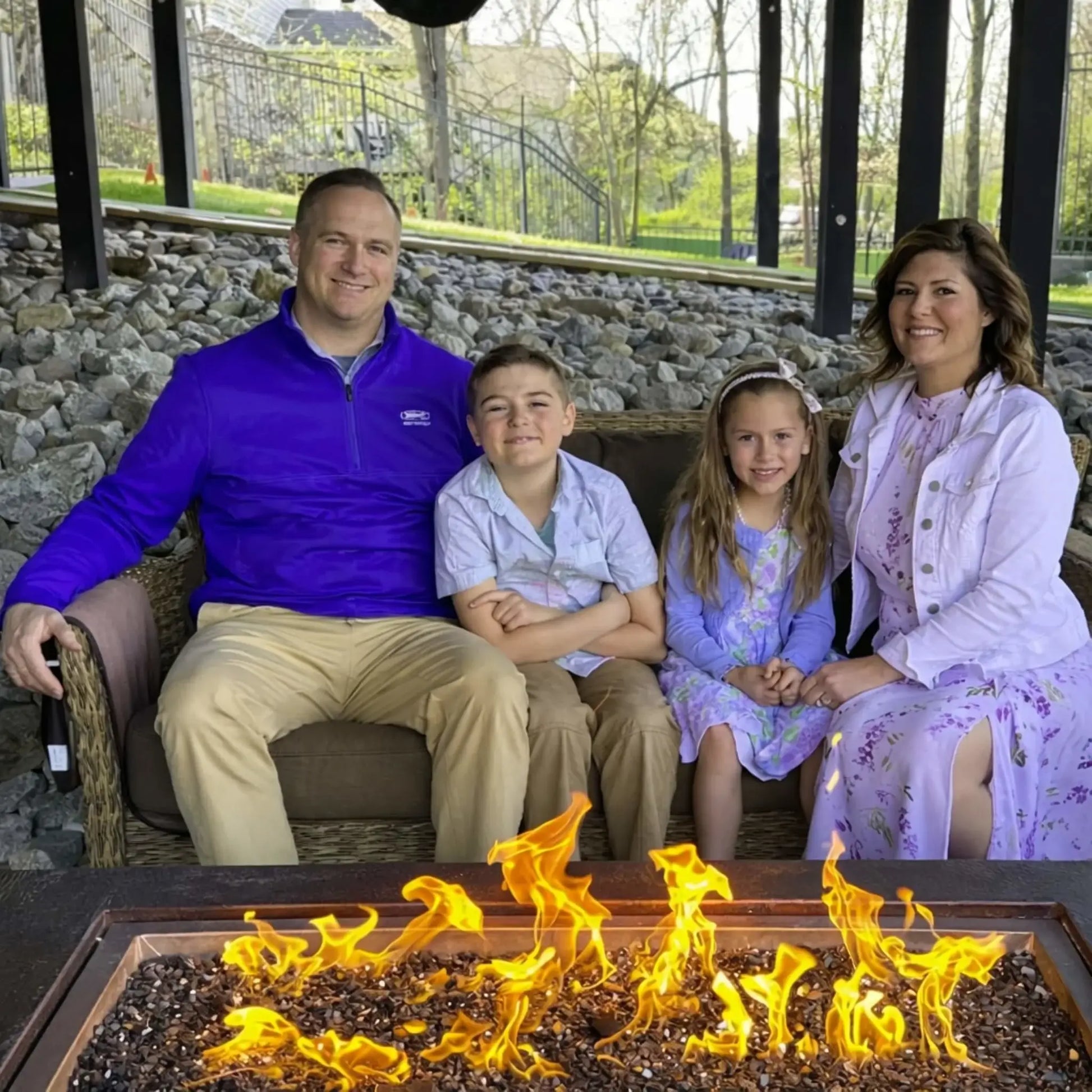 Family enjoying time around the HPC Fire Copper Fire Pit Phoenix H S Rectangle, with a father, mother, and two children seated behind the glowing rectangular fire feature. The flames dance over dark fire media, adding warmth to the covered outdoor living area with stone and wood accents.