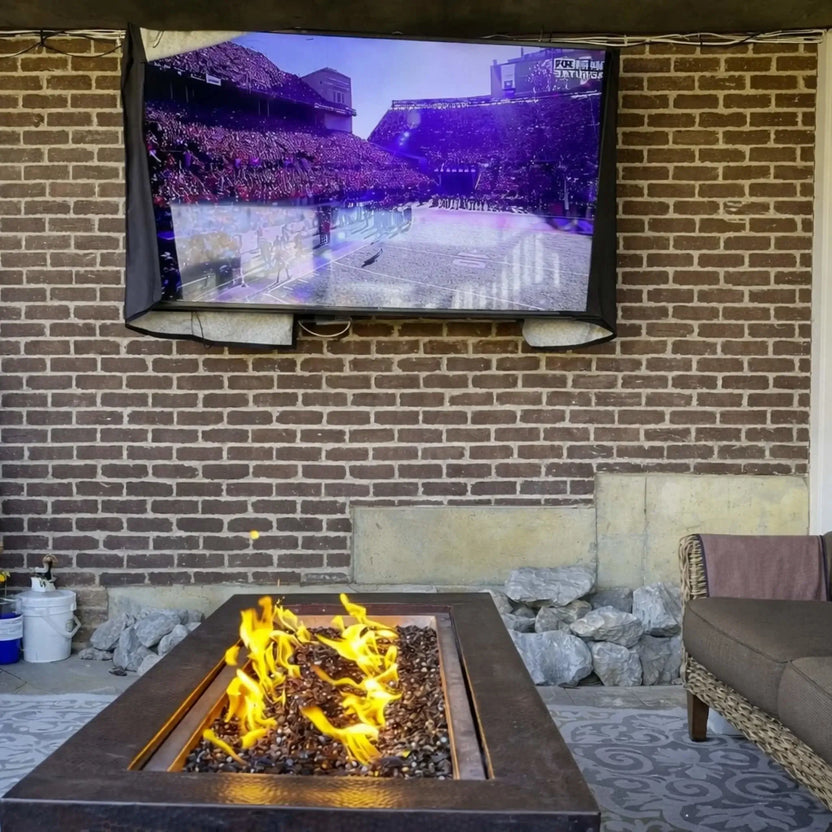 Outdoor entertainment setup featuring the HPC Fire Copper Fire Pit Phoenix H S Rectangle in front of a brick wall with a mounted TV. The rectangular copper fire pit burns brightly with yellow flames over fire glass, creating a relaxed lounge atmosphere with patio seating nearby.