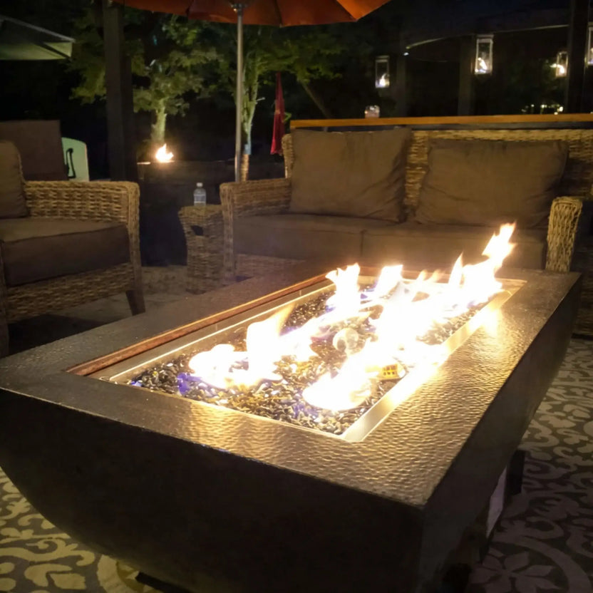 Nighttime patio scene showcasing the HPC Fire Copper Fire Pit Phoenix H S Rectangle surrounded by cushioned wicker seating under a covered structure. The linear flames glow warmly across the fire glass, illuminating the textured copper surface and creating an inviting evening ambiance.