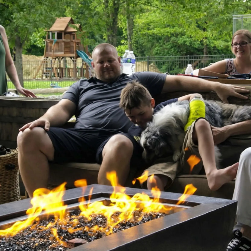 Relaxed outdoor moment featuring the HPC Fire Copper Fire Pit Phoenix H S Rectangle as a family sits nearby with a child and dog on a patio couch. The rectangular fire pit burns steadily in the foreground, with a backyard playset and trees visible in the background.