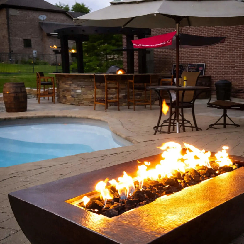 Backyard patio scene featuring the HPC Fire Copper Fire Pit Phoenix Linear Trough installed near a swimming pool, with bright flames rising over dark fire media. The setup includes outdoor seating, a bar area, and umbrella, creating a warm and inviting outdoor entertainment space.