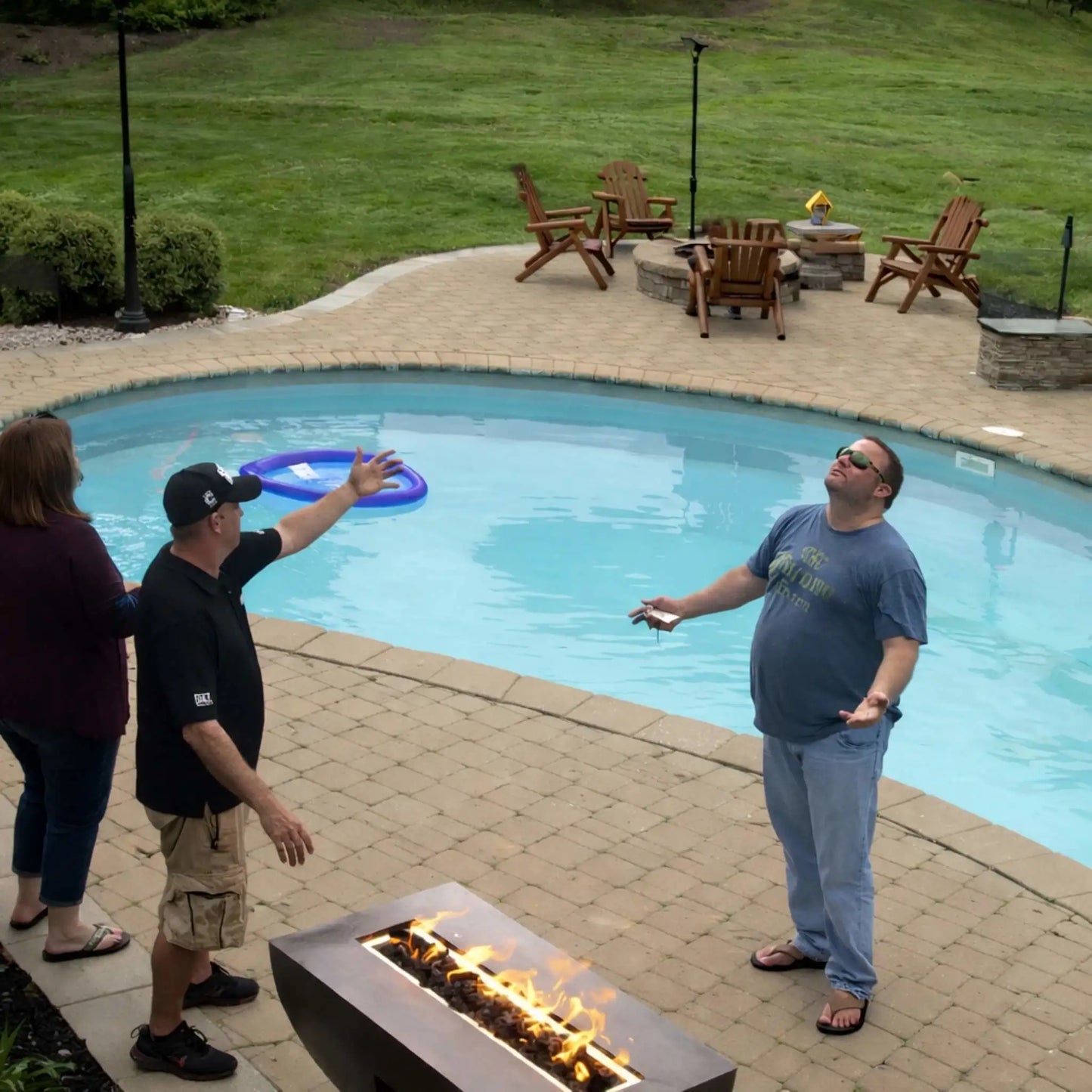 Casual outdoor gathering around the HPC Fire Copper Fire Pit Phoenix Linear Trough beside a backyard pool. Three people stand near the linear fire feature as flames burn steadily, with surrounding patio furniture and open lawn adding to the relaxed residential setting.