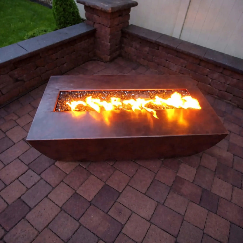 Evening patio installation of the HPC Fire Copper Fire Pit Phoenix Linear Trough, showcasing vibrant flames across the long rectangular burner. The fire feature is set into a paved patio with surrounding brick walls, highlighting its warm glow and modern outdoor design.