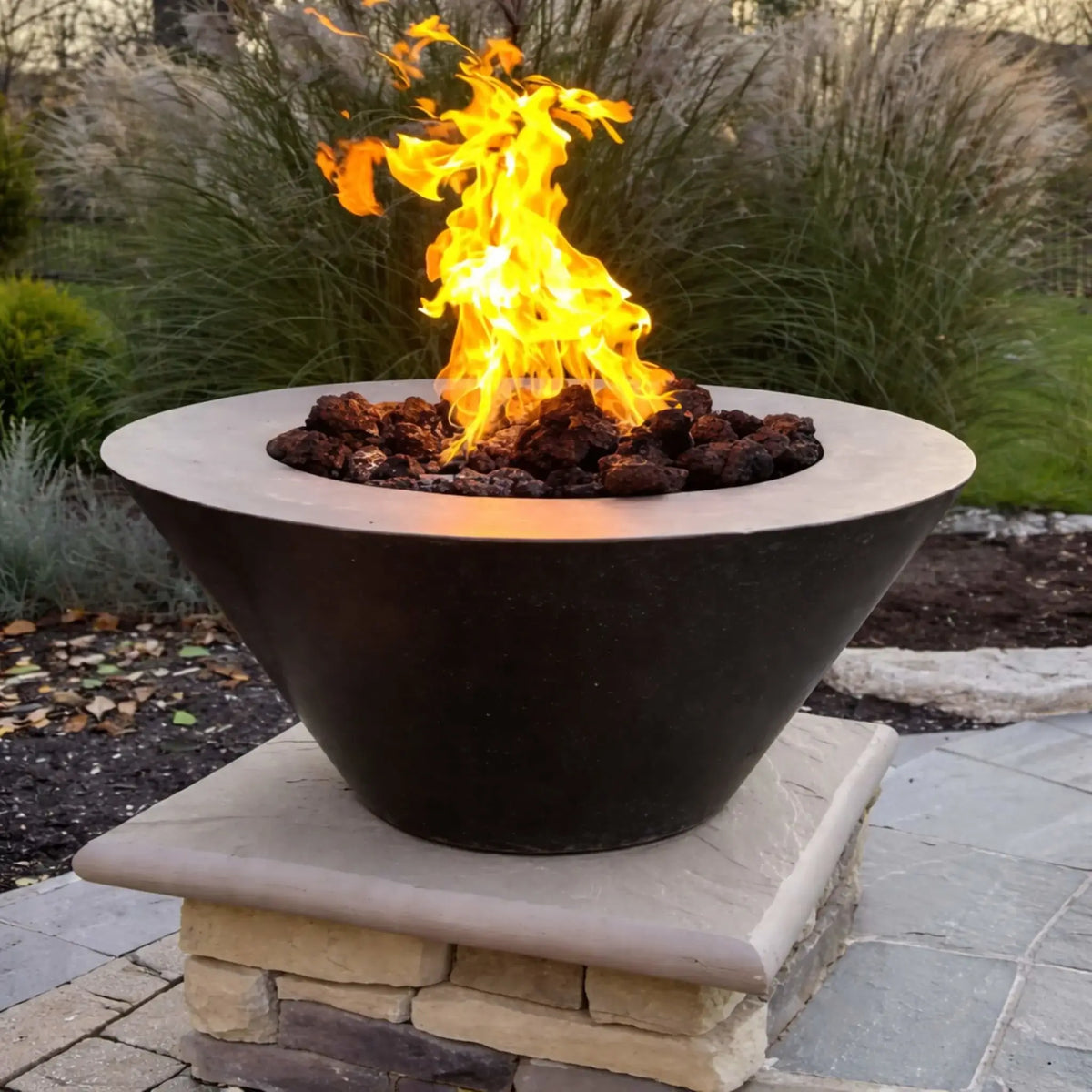 HPC Copper Round Fire Bowl Mesa installed on a stone pedestal base, featuring a bright flame over lava rock, surrounded by ornamental grasses and patio pavers, emphasizing the clean circular design and warm copper-toned finish.