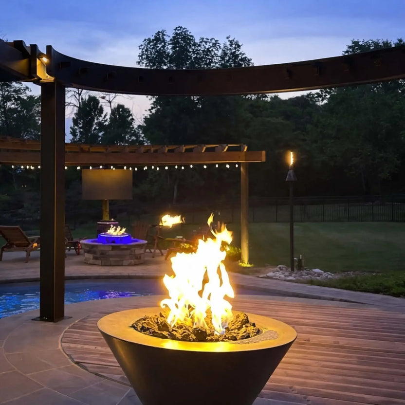 Evening outdoor entertainment area featuring HPC Copper Round Fire Bowl Mesa centered on a raised stone platform under a curved pergola, with ambient lighting, deck seating, and landscaped greenery enhancing the fire bowl’s visual impact.