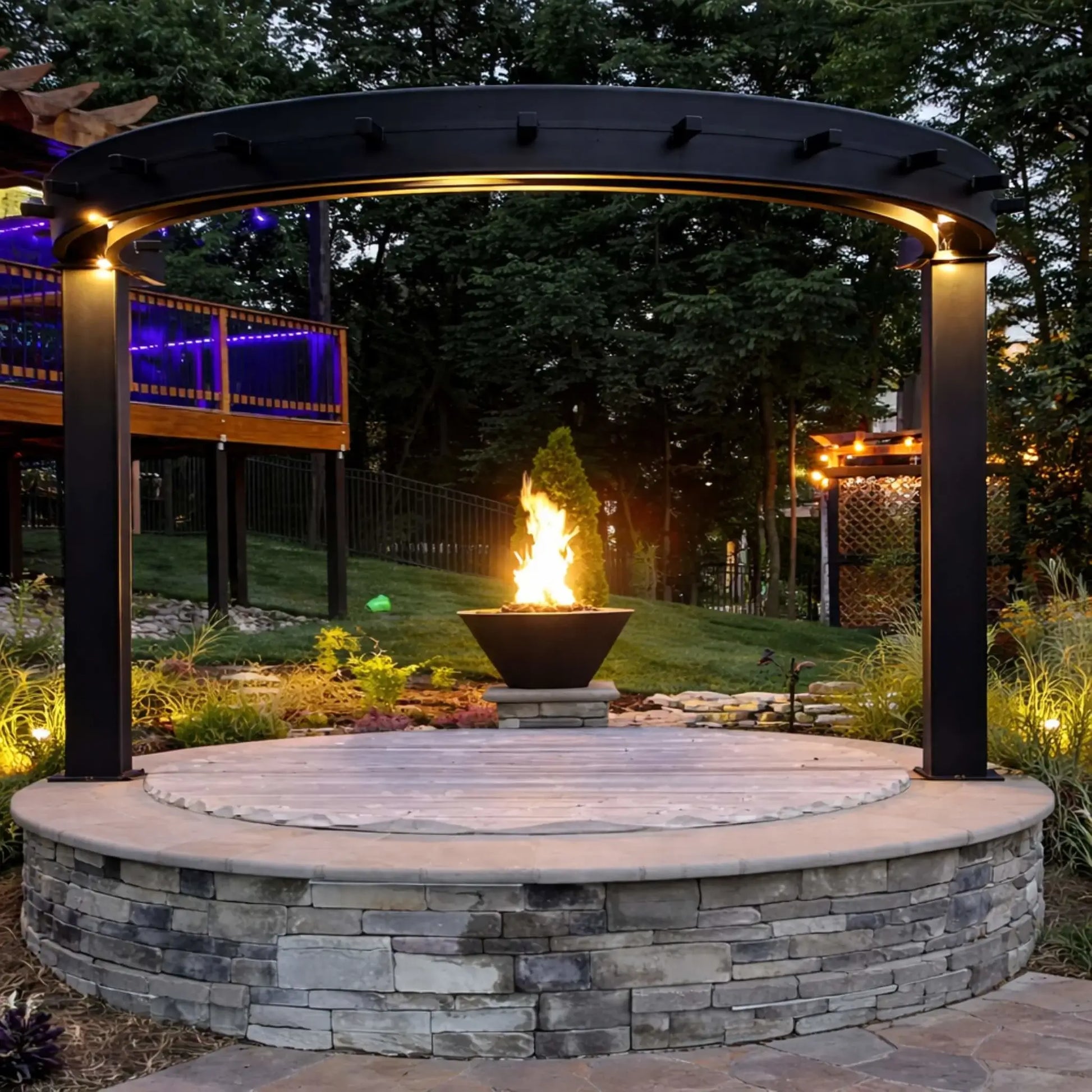 Nighttime backyard setting with HPC Copper Round Fire Bowl Mesa glowing warmly, paired with a secondary illuminated fire feature and seating area, highlighting the bowl’s copper finish and dramatic flame against a dark landscape backdrop.