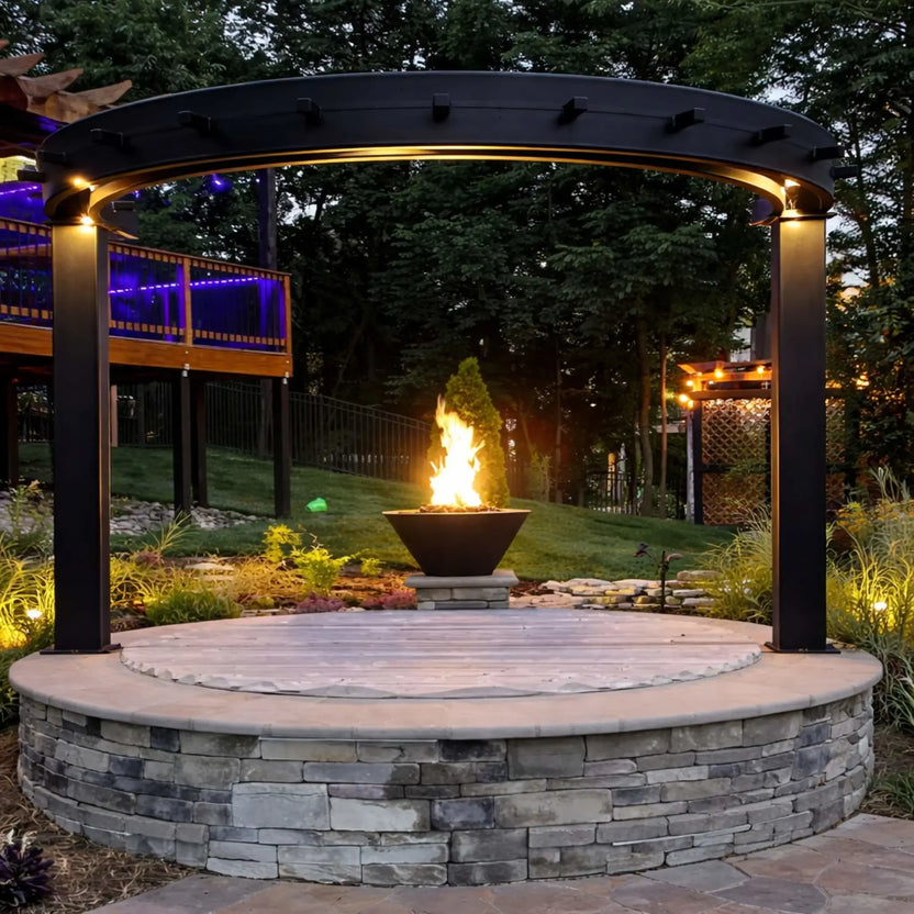 Nighttime backyard setting with HPC Copper Round Fire Bowl Mesa glowing warmly, paired with a secondary illuminated fire feature and seating area, highlighting the bowl’s copper finish and dramatic flame against a dark landscape backdrop.