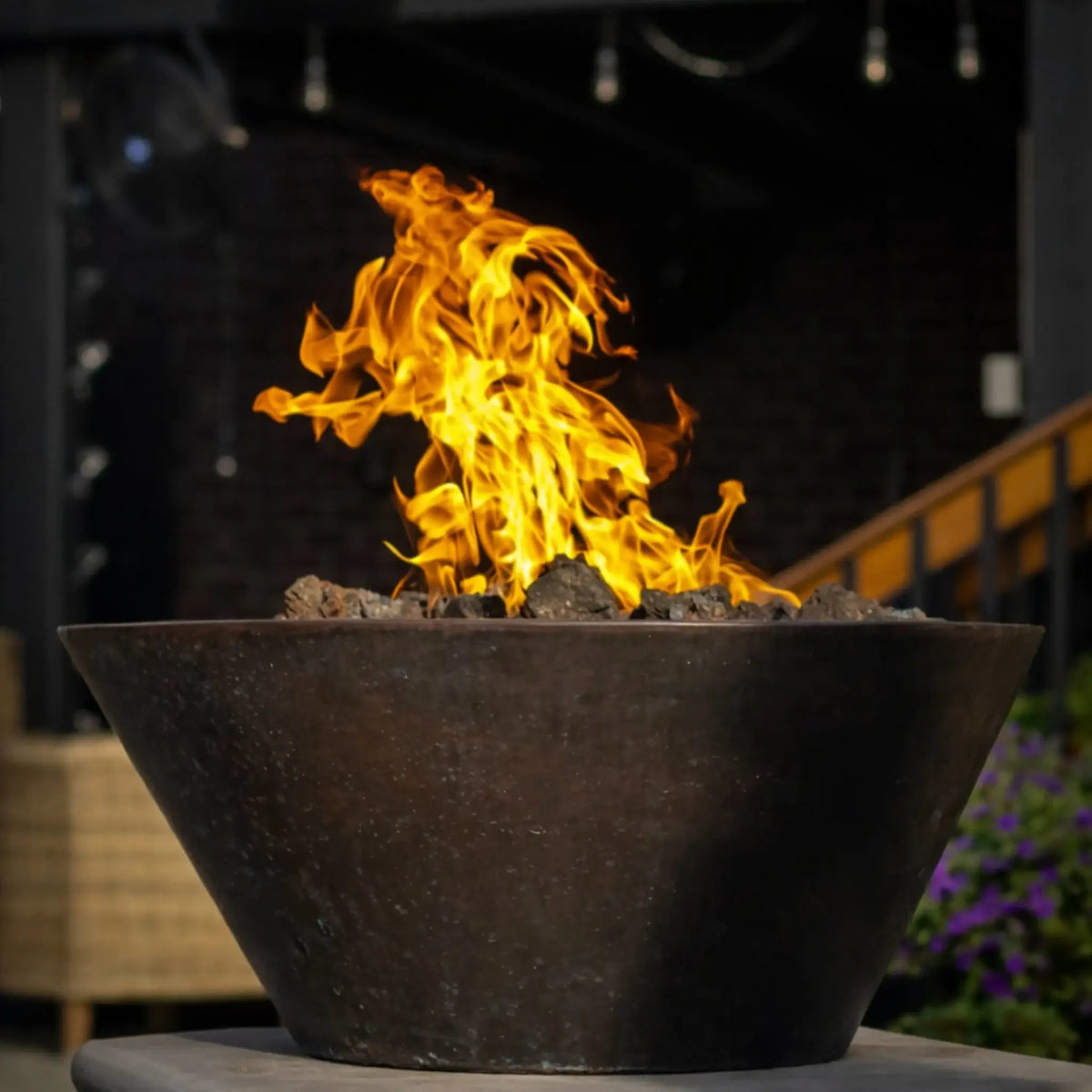 Tight close-up of HPC Copper Round Fire Bowl Mesa flame pattern, showing detailed movement of fire over dark lava rock media, with blurred outdoor structure in background emphasizing flame height and natural gas burner performance.