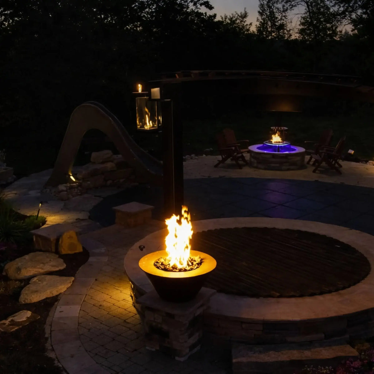 Daytime garden installation of HPC Copper Round Fire Bowl Mesa on a stone pedestal, featuring a low-profile flame, manicured greenery, lattice privacy wall, and patio setting, showcasing the bowl as a decorative outdoor fire feature.