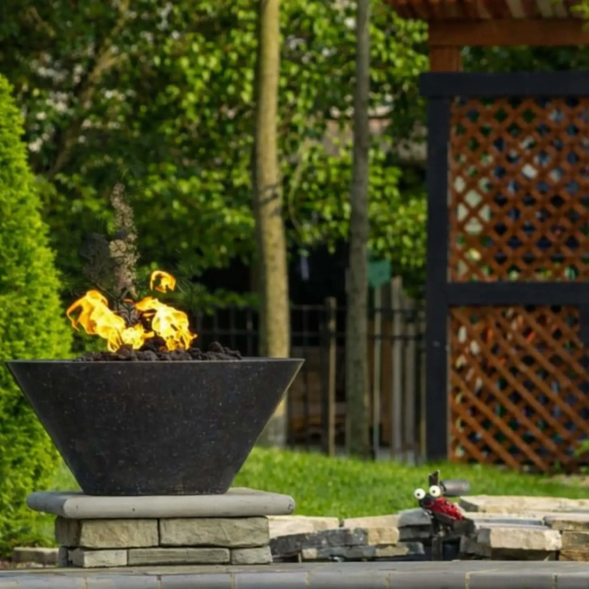 HPC Copper Round Fire Bowl Mesa Lifestyle 8 image showing a dark copper fire bowl with a low, natural gas flame burning over lava rock, positioned on a stacked stone pedestal in a landscaped backyard. The scene includes lush green grass, trimmed shrubs, tall trees, and a wooden pergola with lattice panels, highlighting the fire bowl as a subtle, modern outdoor focal point.