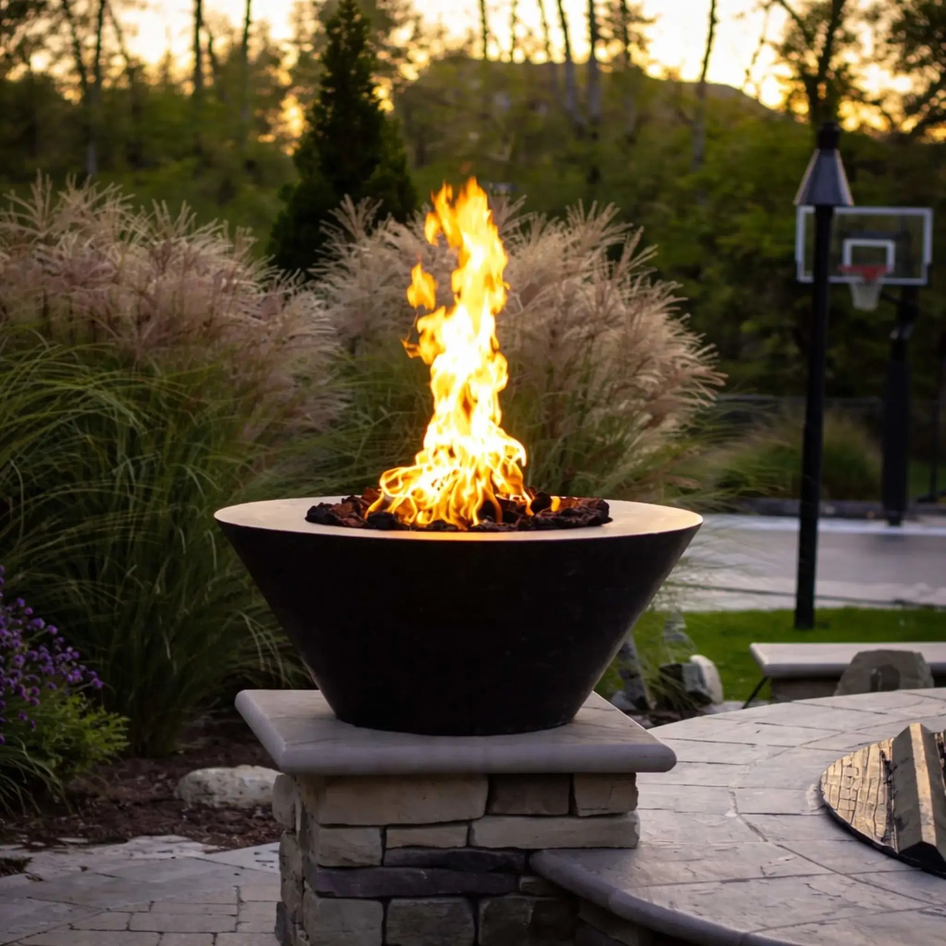 HPC Copper Round Fire Bowl Mesa Lifestyle 9 image showing a tall natural gas flame rising from lava rock inside a dark copper fire bowl, mounted on a stone pedestal beside a curved patio seating wall, surrounded by ornamental grasses and a backyard basketball hoop at sunset.