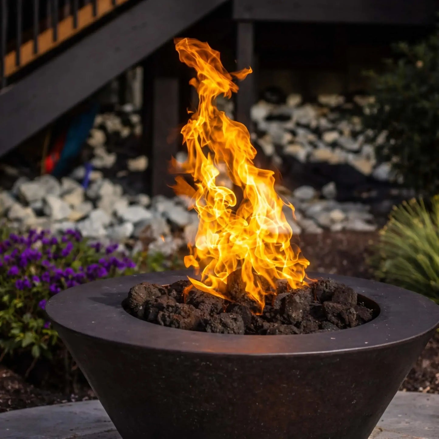 Close-up lifestyle image of HPC Copper Round Fire Bowl Mesa showcasing a vibrant natural gas flame rising above lava rock media, set in a landscaped backyard with flowers and stone accents, highlighting the bowl’s dark copper patina finish.