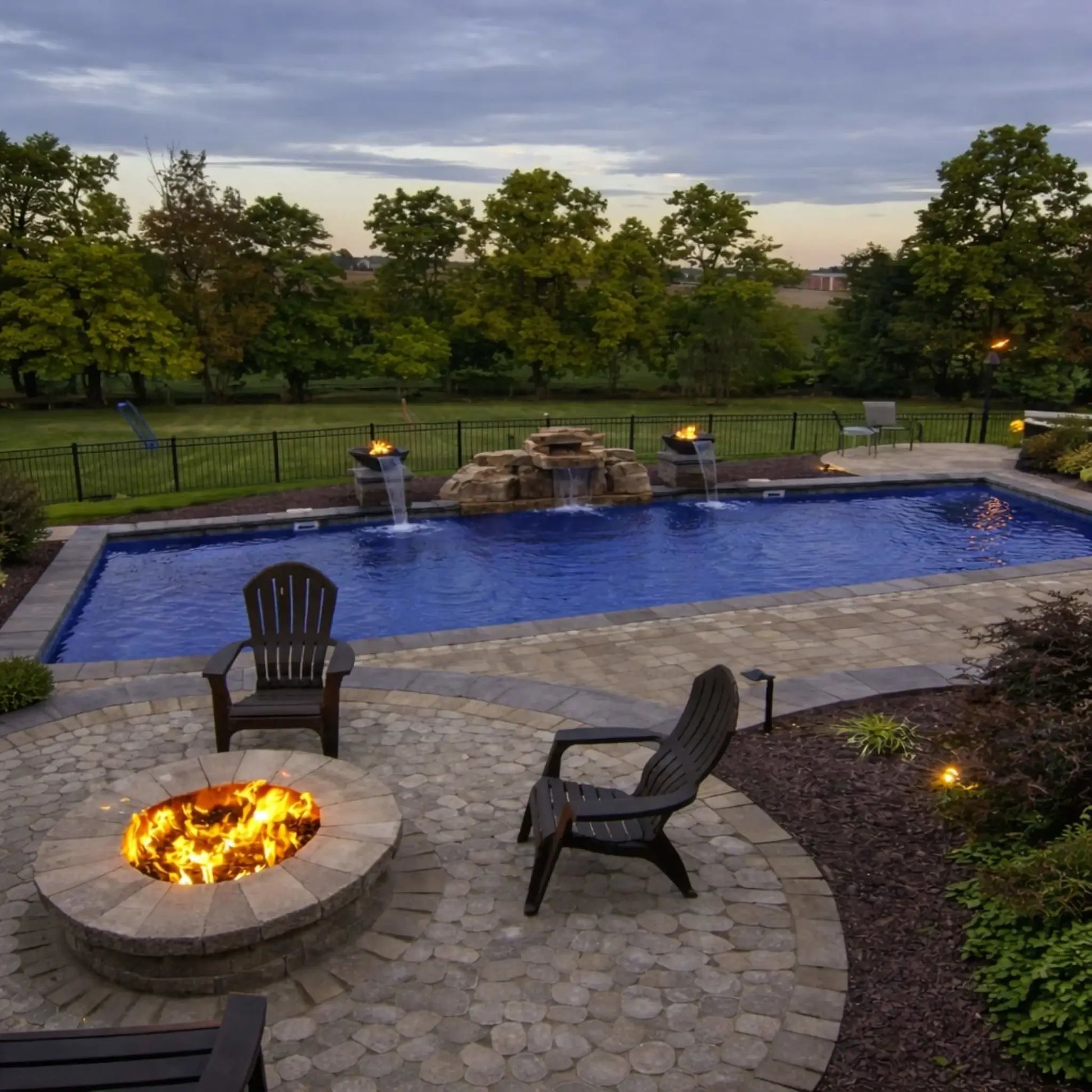 Wide backyard lifestyle shot featuring a luxury poolscape with multiple copper fire and water bowls from the HPC Tempe series installed along the pool edge. Flames rise above each bowl while water cascades into the pool, complemented by a central stone waterfall, paver patio, and Adirondack chairs surrounding a separate fire pit in the foreground.