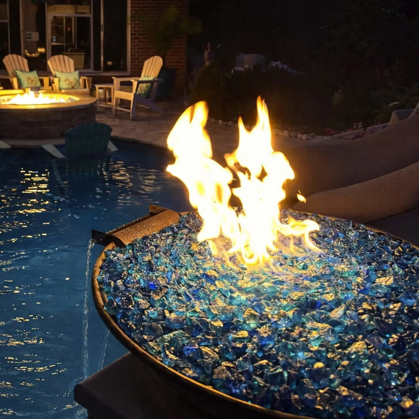 Close-up evening view of HPC Tempe copper fire and water bowl filled with blue fire glass, where bright flames dance above the glass media while water gently pours from the scupper into a swimming pool. The warm glow of the fire contrasts with the cool blue tones of the water, creating a high-end resort-style ambiance.