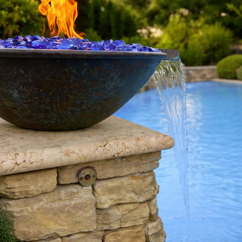 Daytime close-up of HPC Tempe copper fire bowl mounted on a stone column beside a pool, featuring vivid blue fire glass and an active flame. Water flows smoothly from the integrated scupper into the pool below, highlighting the dual fire and water functionality against a landscaped backyard with greenery and stonework.