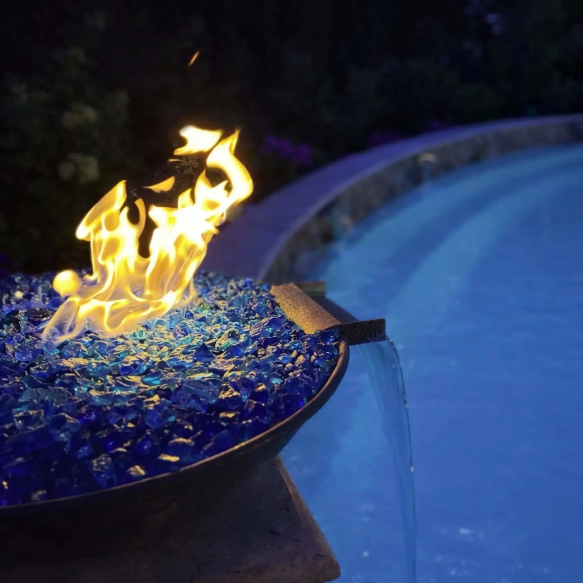 Evening close-up of HPC Tempe copper fire and water bowl glowing with bright flames over reflective blue fire glass, positioned beside a softly lit pool. The scupper releases a steady stream of water, while the darkened surroundings and shimmering reflections emphasize the dramatic fire and water combination in a modern outdoor setting.