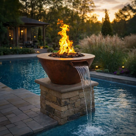 HPC Copper Round Mesa Fire and FireWater water lifestyle 1.0 image showing a hammered copper fire bowl with a strong natural gas flame burning above lava rock, paired with a built-in water scupper spilling a smooth stream into a swimming pool. The fire feature sits on a stacked stone pedestal beside a paver patio, surrounded by ornamental grasses and a luxury backyard setting at sunset.