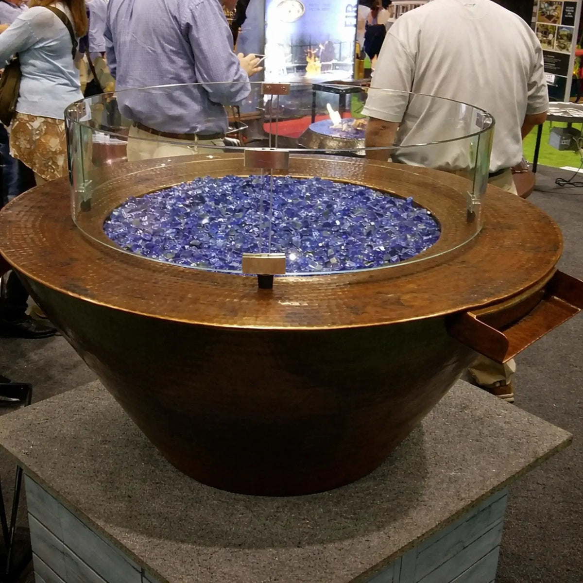 HPC Copper Round Mesa Fire and FireWater water lifestyle 1 image displaying a copper fire bowl filled with blue fire glass and enclosed by a glass wind guard, set in a trade show environment with people in the background, highlighting the fire and water feature design.