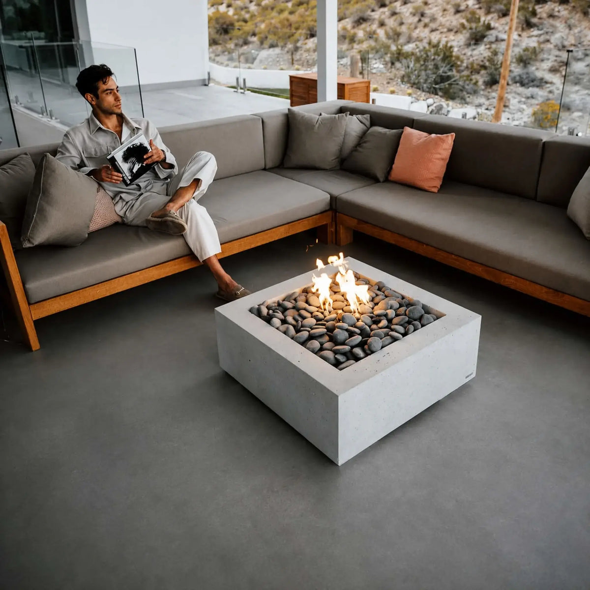 HPC Fire Hearth Products Controls Dekko GFRC Concrete Alea 32 x 32 Fire Pit showcased in a luxury covered outdoor living space. A modern square concrete fire pit filled with smooth gray stones burns at the center of a large sectional sofa while a man relaxes reading nearby. The Alea fire pit serves as a contemporary focal point, blending warmth, comfort, and modern architectural design against a scenic desert backdrop.