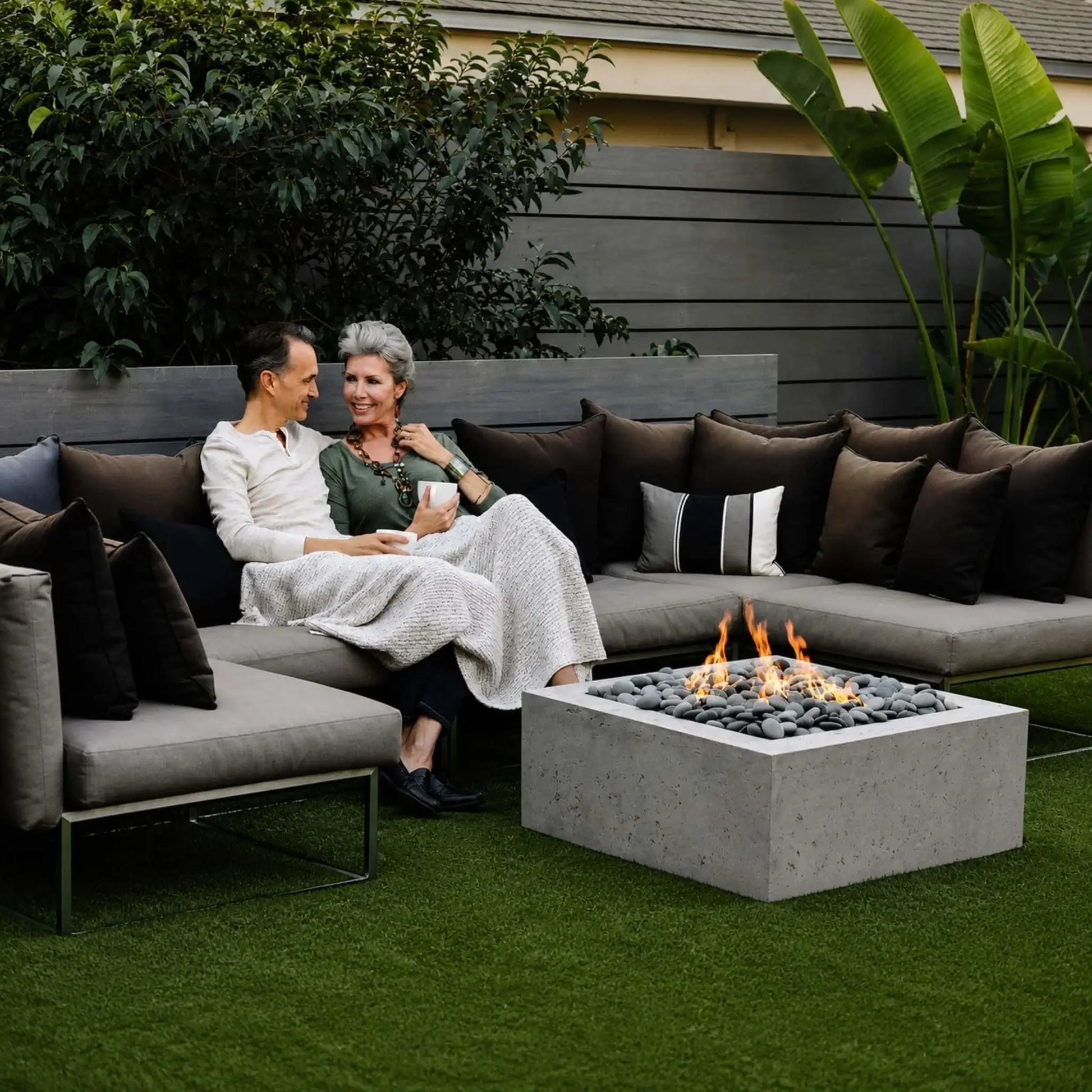 HPC Fire Hearth Products Controls Dekko GFRC Concrete Alea 32 x 32 Fire Pit featured in a stylish backyard lounge setting. The square GFRC concrete fire pit is surrounded by sectional seating and decorative pillows, with flames rising through polished gray stones. A couple relaxes beside the fire, highlighting the Alea fire pit as an inviting centerpiece for outdoor entertaining, conversation, and year-round patio enjoyment.