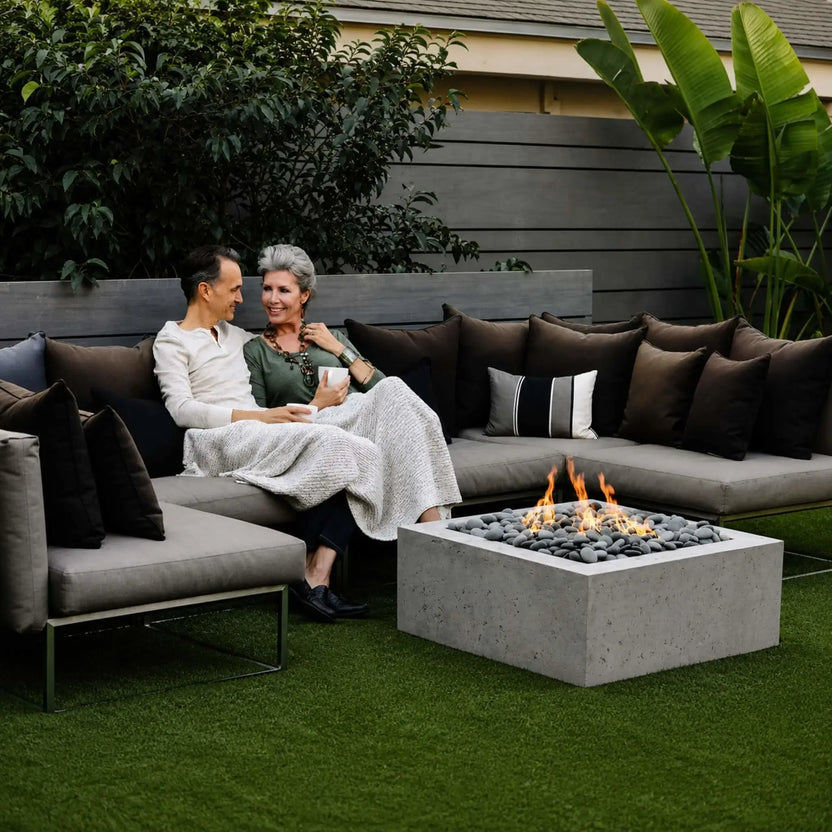 HPC Fire Hearth Products Controls Dekko GFRC Concrete Alea 32 x 32 Fire Pit featured in a stylish backyard lounge setting. The square GFRC concrete fire pit is surrounded by sectional seating and decorative pillows, with flames rising through polished gray stones. A couple relaxes beside the fire, highlighting the Alea fire pit as an inviting centerpiece for outdoor entertaining, conversation, and year-round patio enjoyment.
