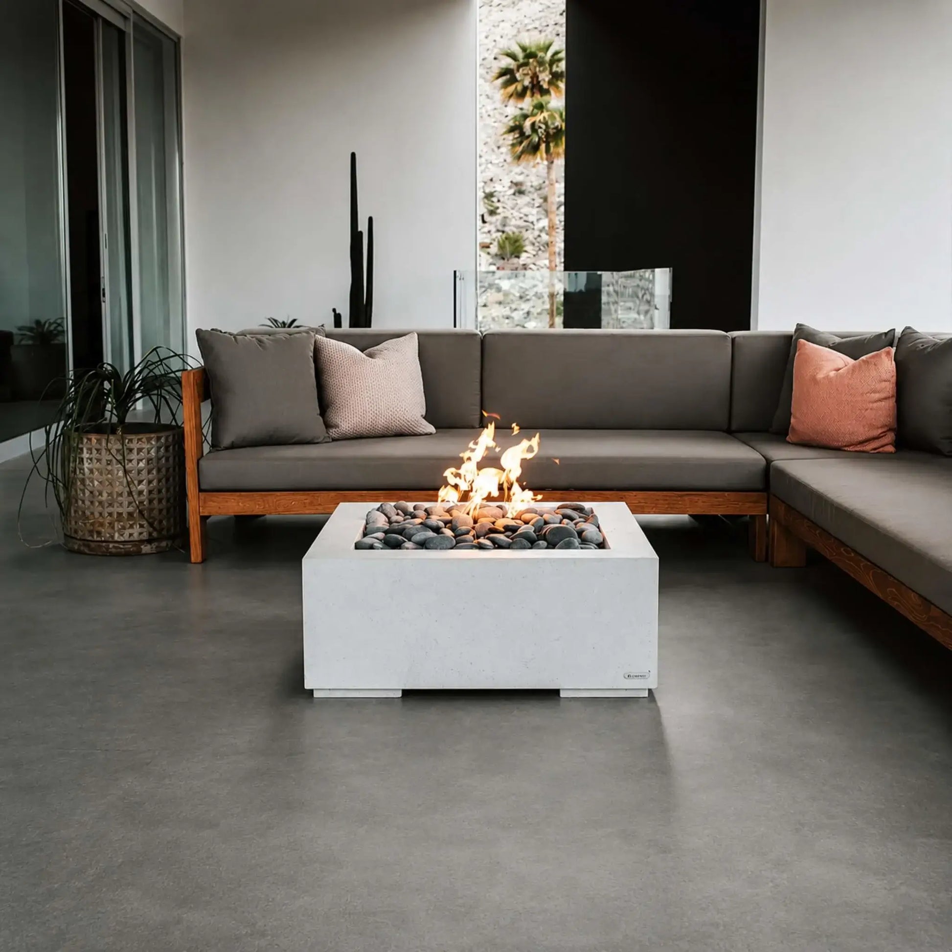 HPC Fire Hearth Products Controls Dekko GFRC Concrete Alea 32 x 32 Fire Pit positioned in a modern covered patio with contemporary sectional seating. Bright flames emerge from decorative river stones inside the square concrete fire pit, creating a warm atmosphere. The Alea fire pit complements clean architectural lines, outdoor furnishings, and upscale residential living spaces designed for relaxation and entertaining.