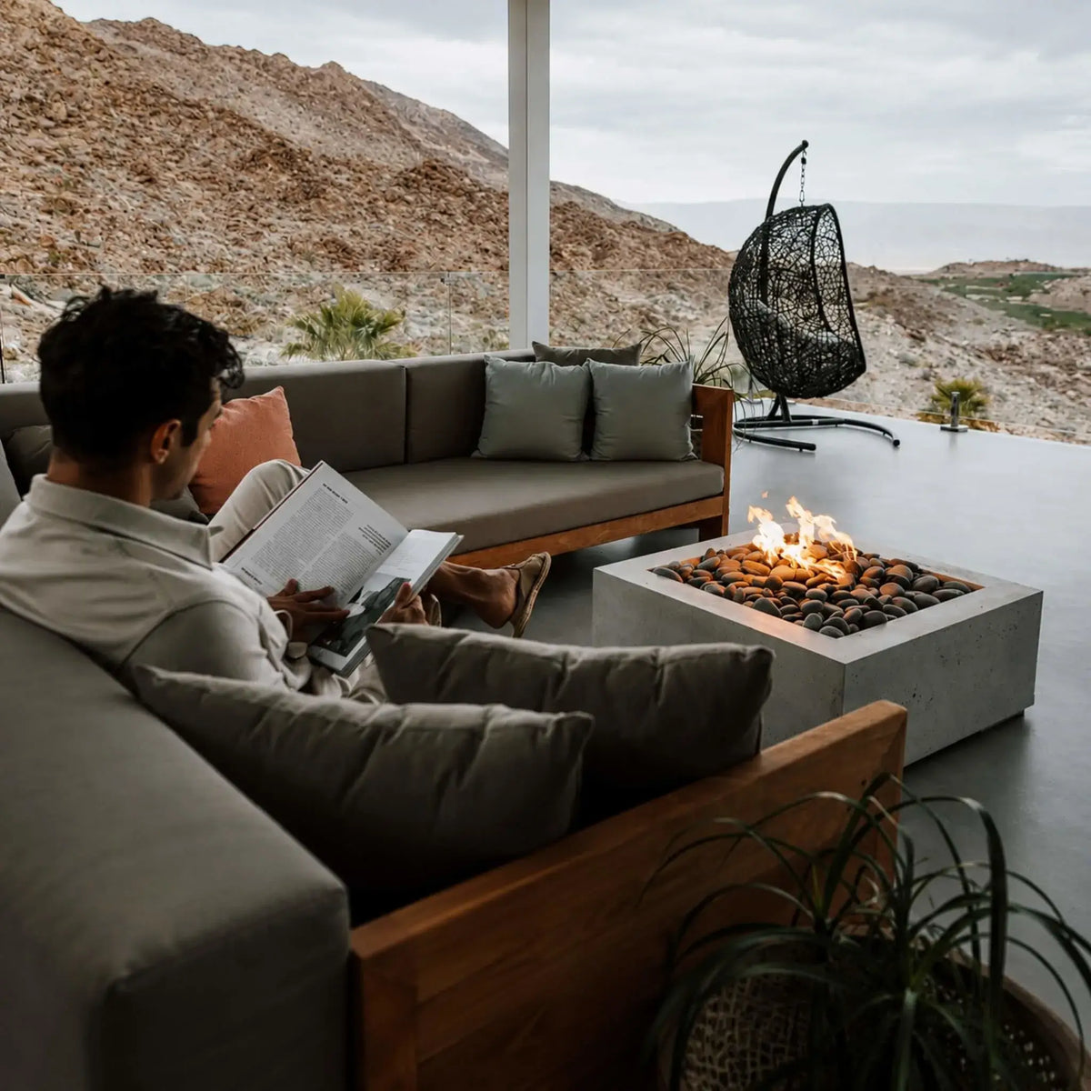 HPC Fire Hearth Products Controls Dekko GFRC Concrete Alea 32 x 32 Fire Pit displayed on a luxury desert-view terrace. A person reads comfortably on a cushioned outdoor sectional while flames glow from the stone-filled concrete fire pit. Surrounded by mountain scenery, modern furnishings, and a hanging chair, the Alea GFRC fire pit creates an elegant outdoor retreat for relaxation and evening gatherings.