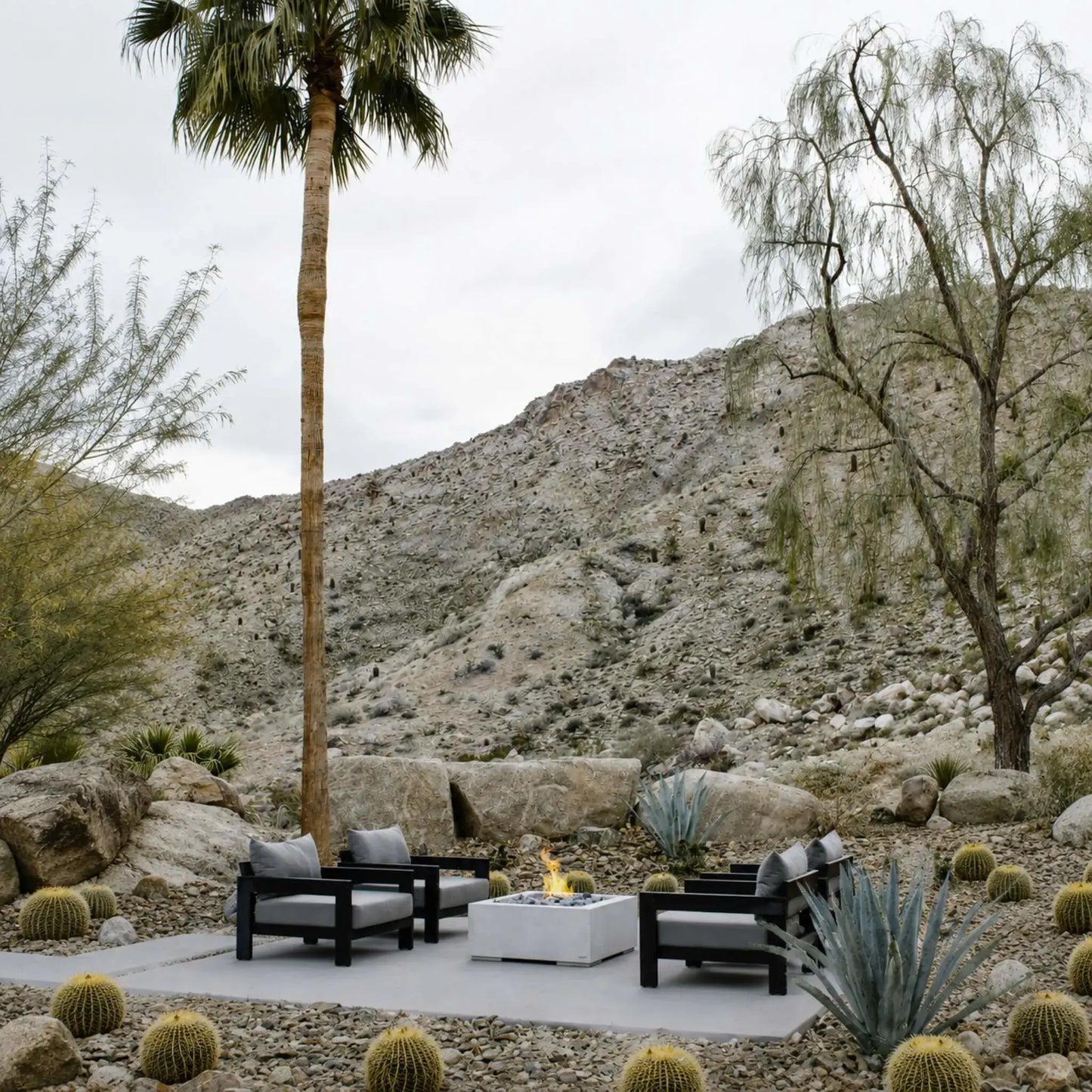 HPC Fire Hearth Products Controls Dekko GFRC Concrete Alea 32 x 32 Fire Pit installed in a landscaped desert outdoor living area. The square concrete fire pit sits on a patio surrounded by lounge chairs, palm trees, cacti, natural boulders, and mountain views. A small flame burns at the center, showcasing the Alea fire pit as a stylish and functional gathering space within a contemporary desert-inspired landscape design.