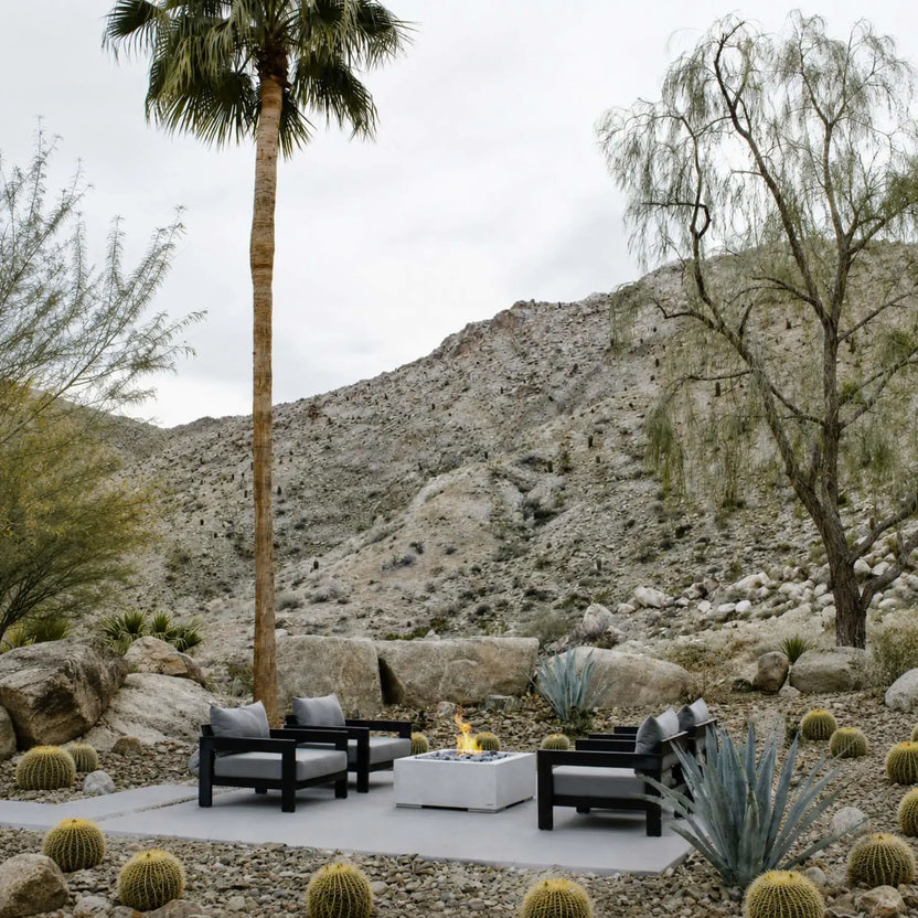 HPC Fire Hearth Products Controls Dekko GFRC Concrete Alea 32 x 32 Fire Pit installed in a landscaped desert outdoor living area. The square concrete fire pit sits on a patio surrounded by lounge chairs, palm trees, cacti, natural boulders, and mountain views. A small flame burns at the center, showcasing the Alea fire pit as a stylish and functional gathering space within a contemporary desert-inspired landscape design.