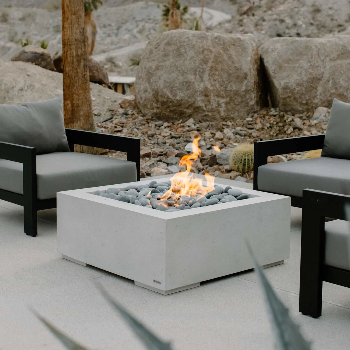 HPC Fire Hearth Products Controls Dekko GFRC Concrete Alea 32 x 32 Fire Pit featured in a modern outdoor lounge setting with black-framed patio chairs and gray cushions. The square GFRC concrete fire pit showcases smooth river rock media surrounding a natural gas flame, creating a clean and contemporary focal point. Set against a desert-inspired landscape with large boulders and native plants, the Alea 32 x 32 fire pit blends modern design, warmth, and durable outdoor performance.