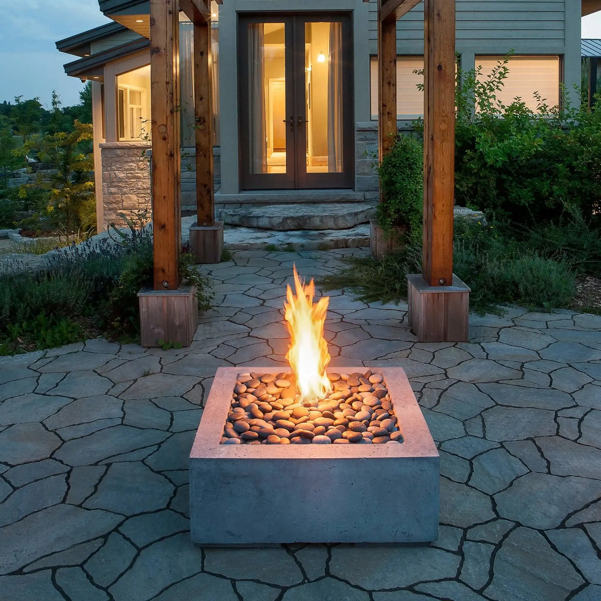HPC Fire Hearth Products Controls Dekko GFRC Concrete Alea 32 x 32 Fire Pit installed on a natural stone patio at the entrance of a luxury home. The square concrete fire feature is filled with polished river stones and displays a vibrant flame rising from the center. Framed by timber posts, stone pathways, and illuminated glass entry doors, this Alea 32 x 32 fire pit highlights contemporary outdoor living and elegant architectural design for residential landscapes.