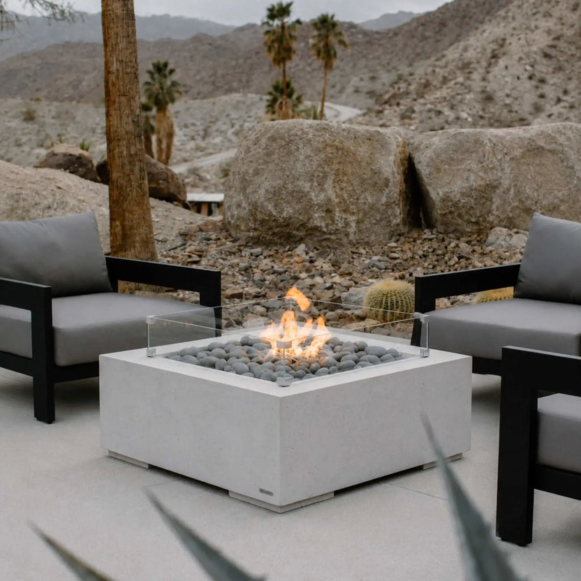 Lifestyle image of the HPC Fire Hearth Products Controls Dekko GFRC Concrete Alea 32 x 32 Fire Pit with tempered glass wind guard installed on a luxury outdoor patio. The white GFRC concrete fire pit features gray fire stones and active flames, surrounded by modern lounge chairs. Desert mountains, boulders, palm trees, and natural landscaping create a sophisticated resort-style outdoor living setting.