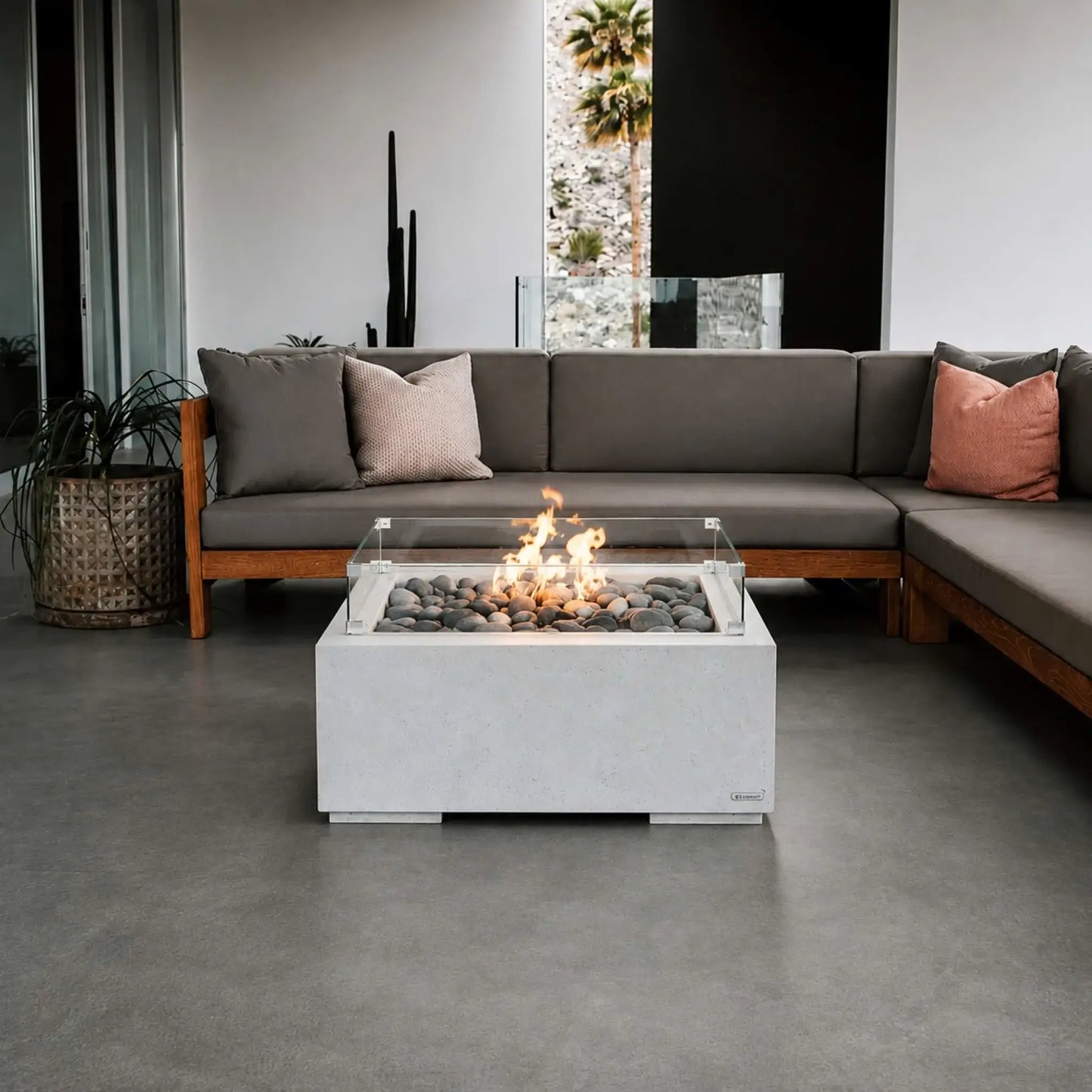 HPC Fire Hearth Products Controls Dekko GFRC Concrete Alea 32 x 32 Fire Pit with tempered glass wind guard featured in a contemporary covered patio lounge. The white GFRC concrete fire pit burns over smooth gray fire stones and is positioned in front of a large sectional sofa with accent pillows. Clean architectural lines, modern furnishings, and warm flames create an inviting outdoor living atmosphere.