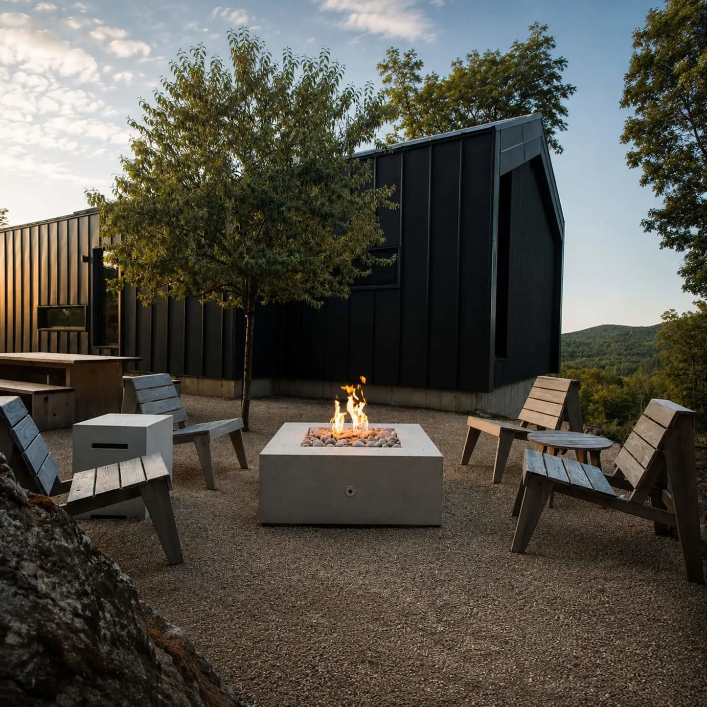HPC Fire Hearth Products Controls Dekko GFRC Concrete Alea 40 x 40 Fire Pit featured in a natural outdoor setting with a woman tending the fire. The square GFRC concrete fire pit is filled with smooth white decorative stones and active flames, positioned on a wood deck surrounded by rugged rock formations and greenery. A modern black lounge chair with a folded blanket completes the cozy, upscale outdoor living atmosphere.