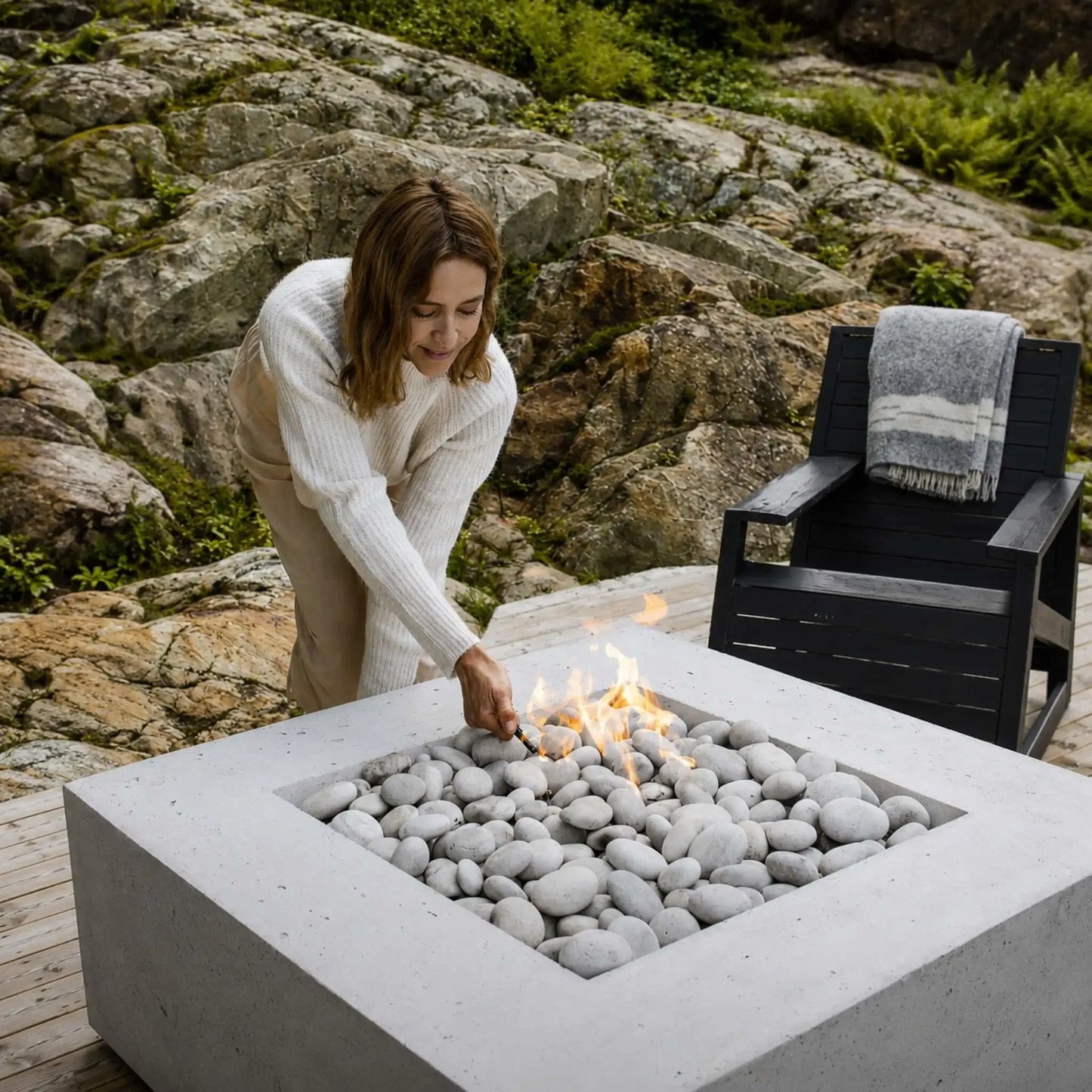 HPC Fire Hearth Products Controls Dekko GFRC Concrete Alea 40 x 40 Fire Pit featured in a natural outdoor setting with a woman tending the fire. The square GFRC concrete fire pit is filled with smooth white decorative stones and active flames, positioned on a wood deck surrounded by rugged rock formations and greenery. A modern black lounge chair with a folded blanket completes the cozy, upscale outdoor living atmosphere.