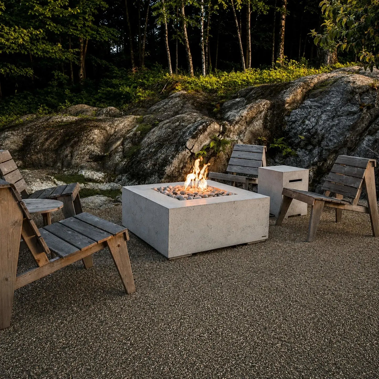 HPC Fire Hearth Products Controls Dekko GFRC Concrete Alea 40 x 40 Fire Pit positioned among rustic outdoor chairs in a woodland-inspired setting. The square concrete fire pit features a bed of smooth decorative stones with natural flames rising above the burner, while surrounding rock formations and mature trees create a warm, inviting retreat for outdoor relaxation.