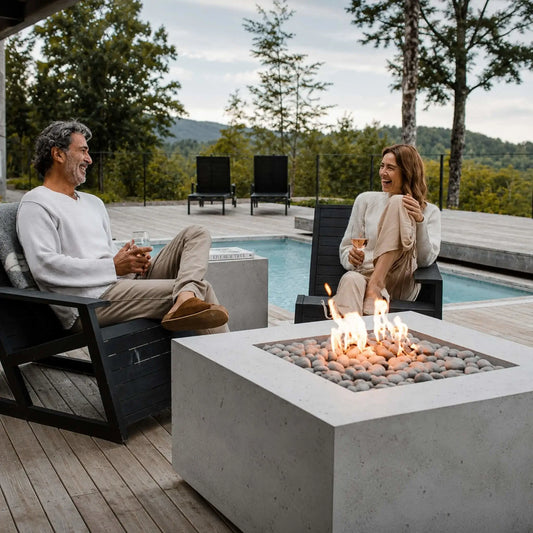HPC Fire Hearth Products Controls Dekko GFRC Concrete Alea 40 x 40 Fire Pit showcased in a premium backyard lounge area overlooking a pool and wooded landscape. A couple relaxes beside the fire pit in contemporary outdoor chairs, enjoying the warmth of the flames above decorative stones. The scene highlights the fire pit’s role as a stylish focal point for outdoor entertaining.