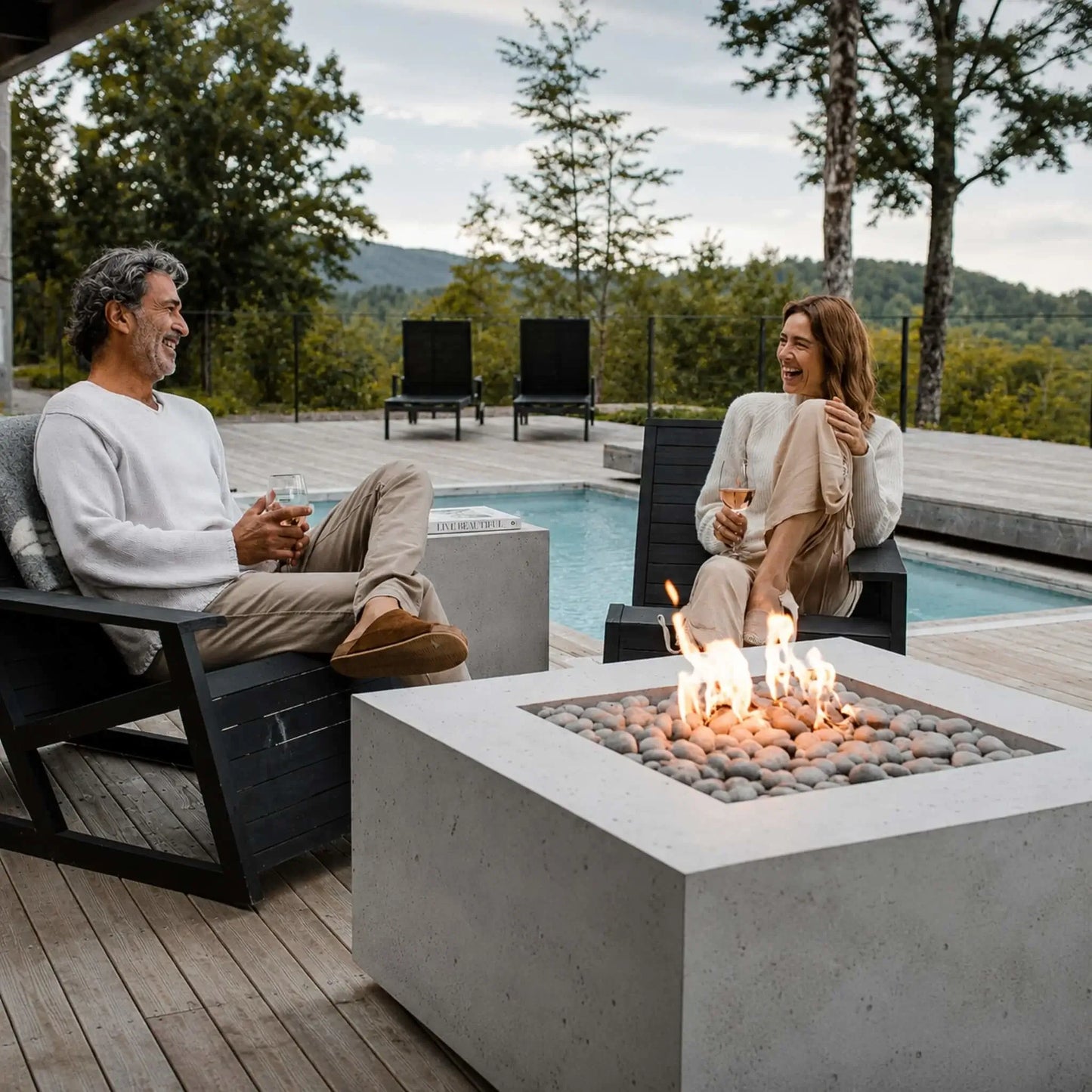HPC Fire Hearth Products Controls Dekko GFRC Concrete Alea 40 x 40 Fire Pit showcased in a premium backyard lounge area overlooking a pool and wooded landscape. A couple relaxes beside the fire pit in contemporary outdoor chairs, enjoying the warmth of the flames above decorative stones. The scene highlights the fire pit’s role as a stylish focal point for outdoor entertaining.