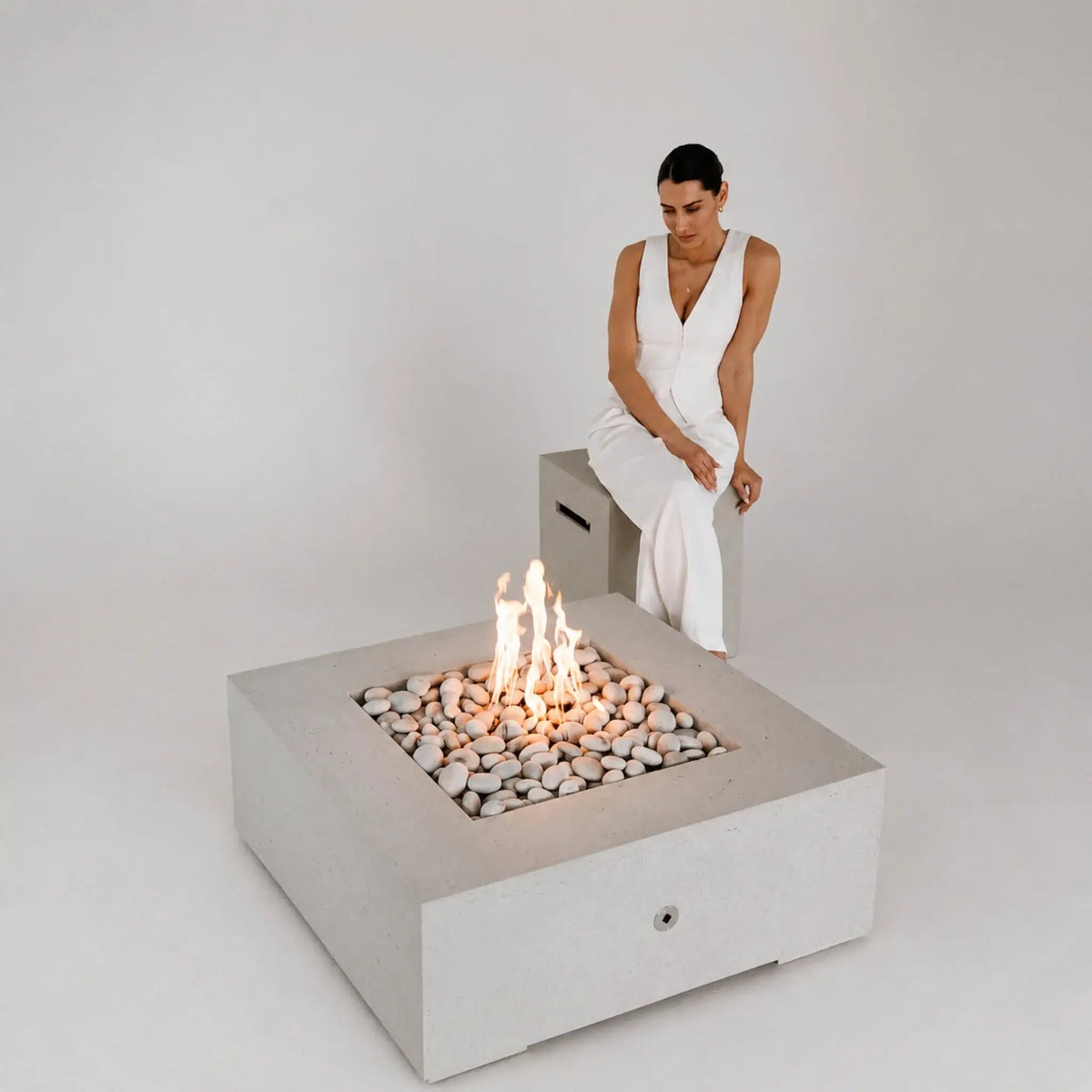 HPC Fire Hearth Products Controls Dekko GFRC Concrete Alea 40 x 40 Fire Pit featured in a minimalist studio setting with a woman seated beside the fire feature. The square GFRC concrete fire pit contains smooth decorative stones and bright flames, highlighting its clean geometric design, contemporary styling, and suitability for modern residential or commercial outdoor environments.