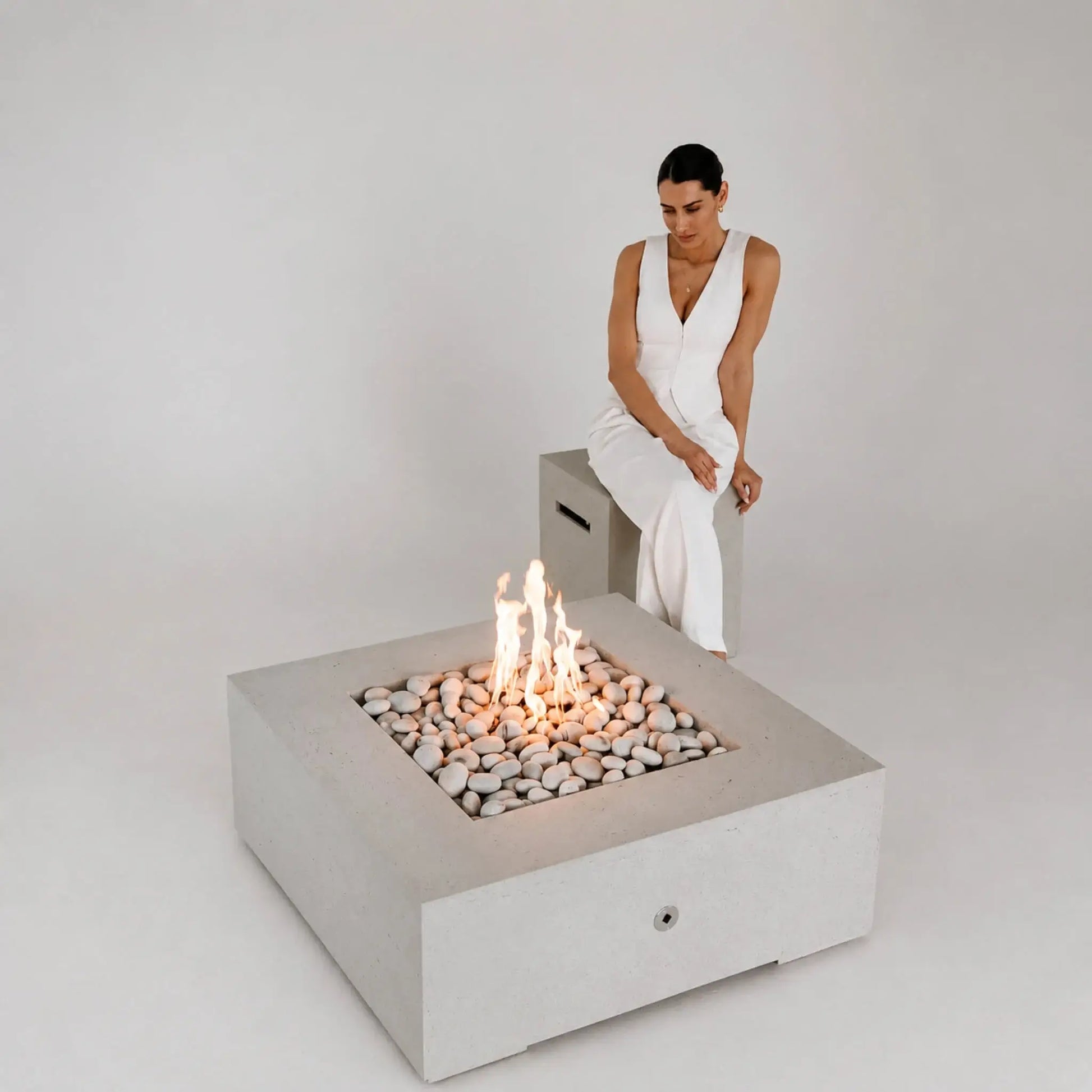 HPC Fire Hearth Products Controls Dekko GFRC Concrete Alea 40 x 40 Fire Pit featured in a minimalist studio setting with a woman seated beside the fire feature. The square GFRC concrete fire pit contains smooth decorative stones and bright flames, highlighting its clean geometric design, contemporary styling, and suitability for modern residential or commercial outdoor environments.