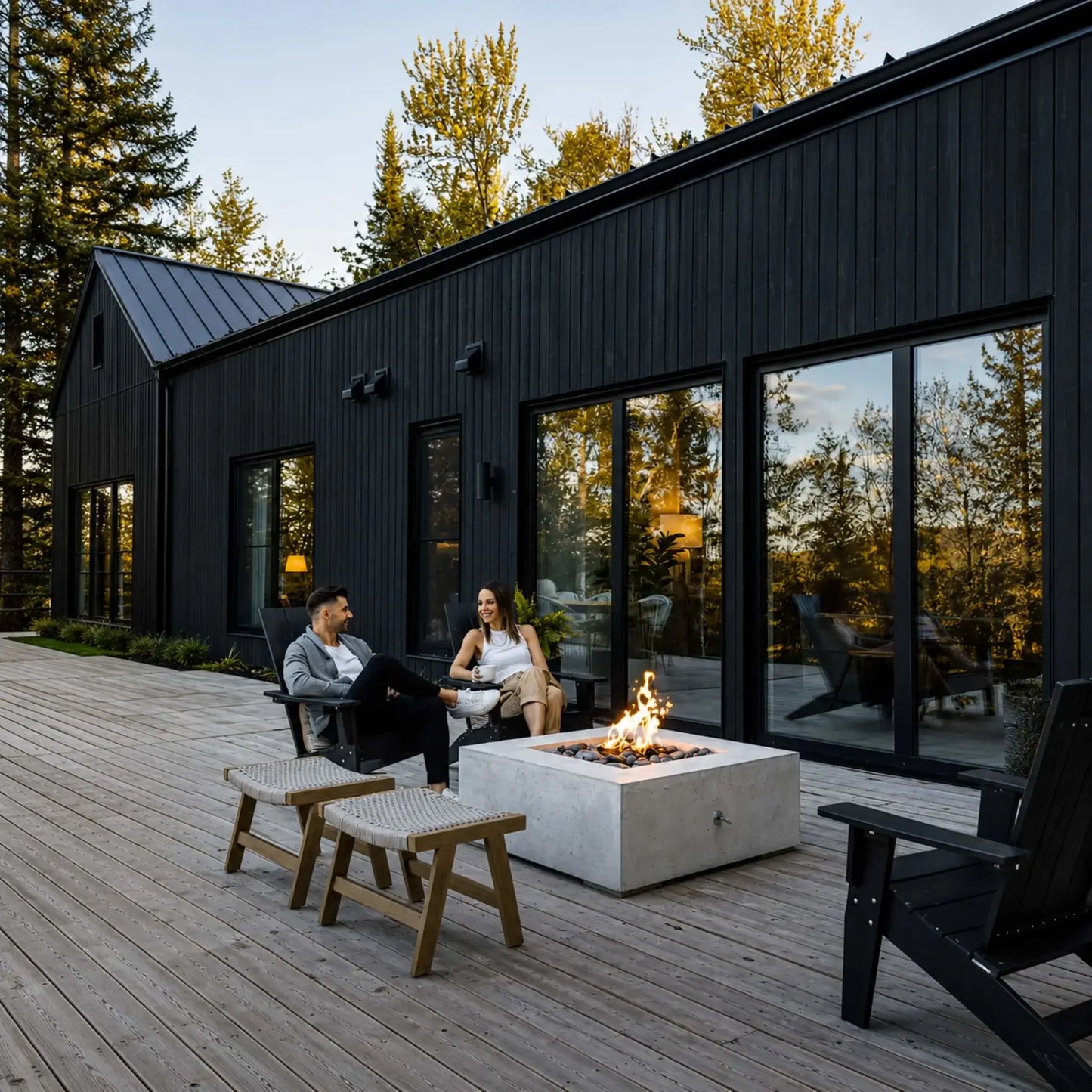 HPC Fire Hearth Products Controls Dekko GFRC Concrete Alea 40 x 40 Fire Pit installed on a spacious wooden deck outside a modern black exterior home. The square concrete fire pit burns with a vibrant flame over decorative stone media while two people relax nearby. Large glass doors, contemporary architecture, and forest views emphasize the upscale outdoor living experience.